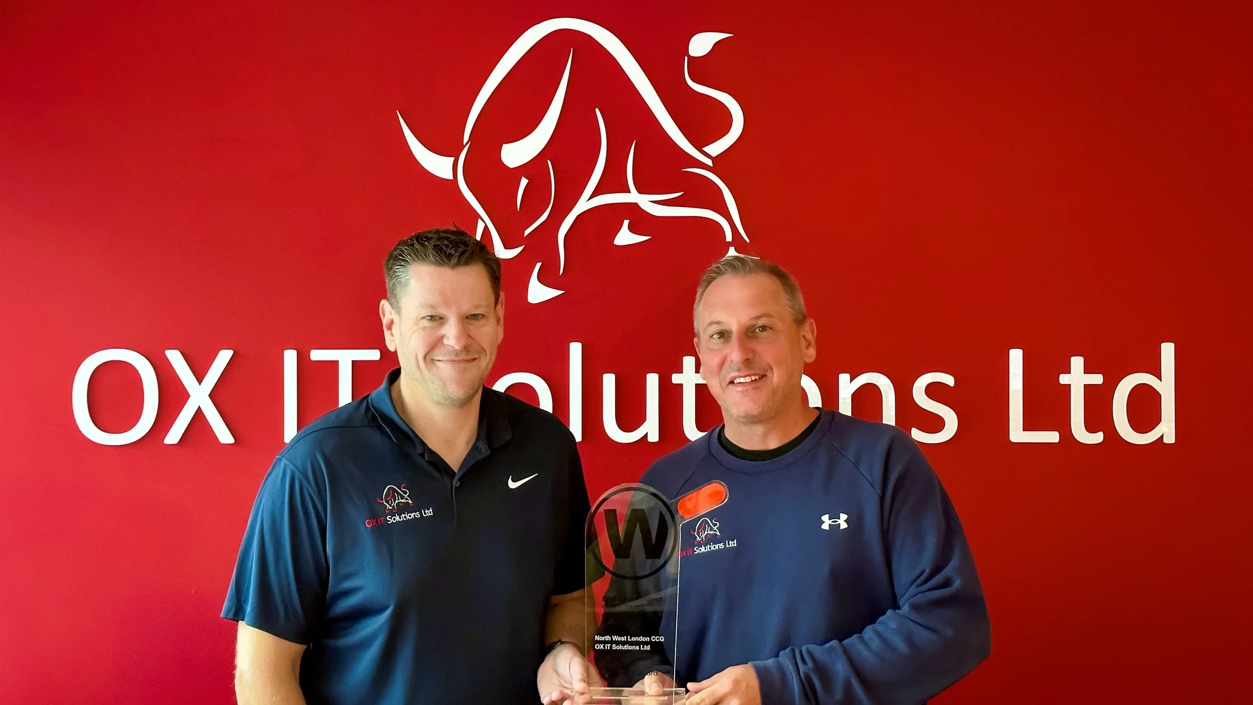 Two men standing side by side in front of a red background with a white bull logo and the text "OX IT Solutions Ltd." The man on the left is smiling and wearing a dark blue polo shirt, while the man on the right is wearing a dark blue Under Armour sh