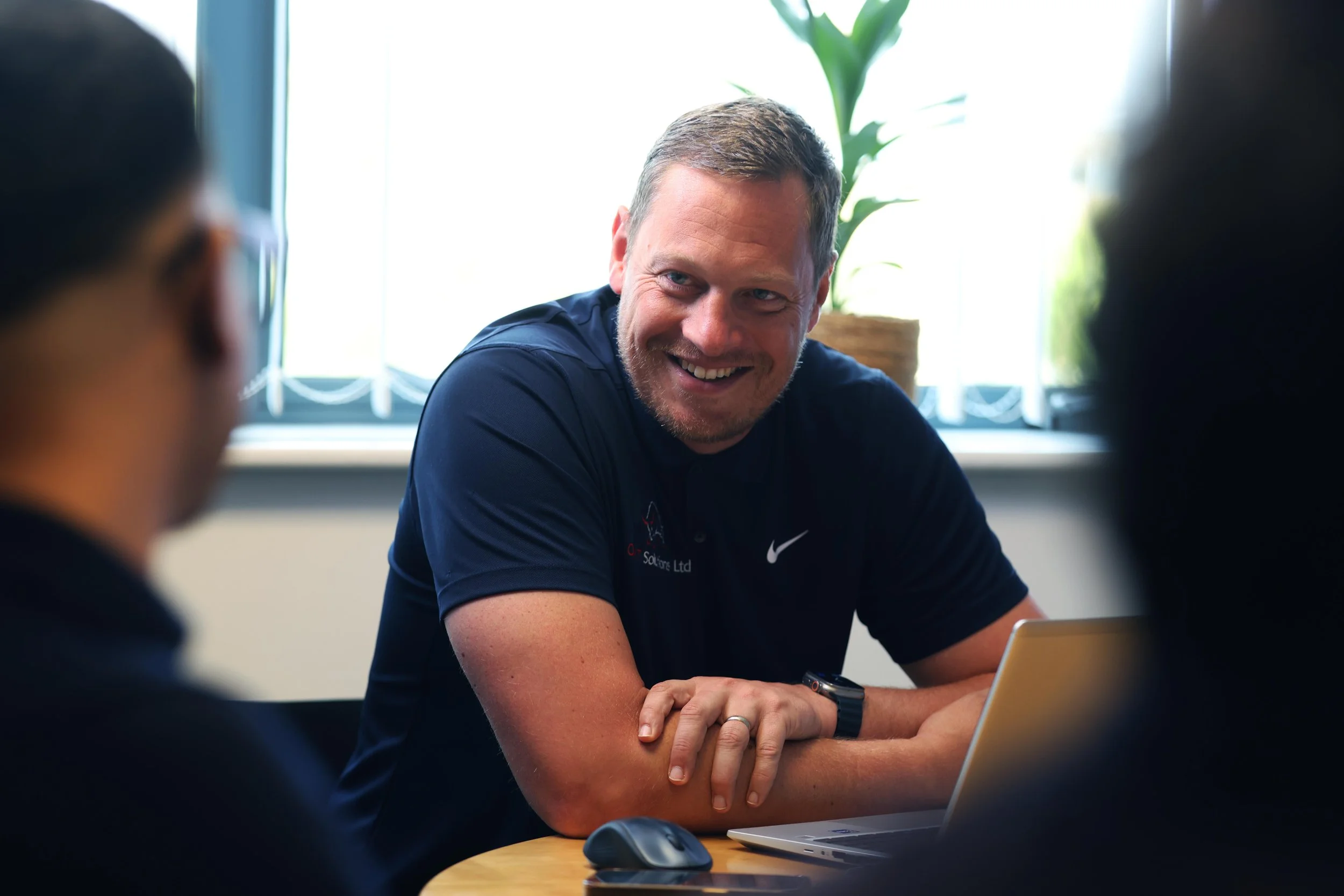 A man with short hair and a beard, wearing a dark polo shirt, is smiling and engaging in a conversation with two other people in an office setting. There is a laptop and a computer mouse on the table in front of him, and a potted plant is visible behind him near a window.