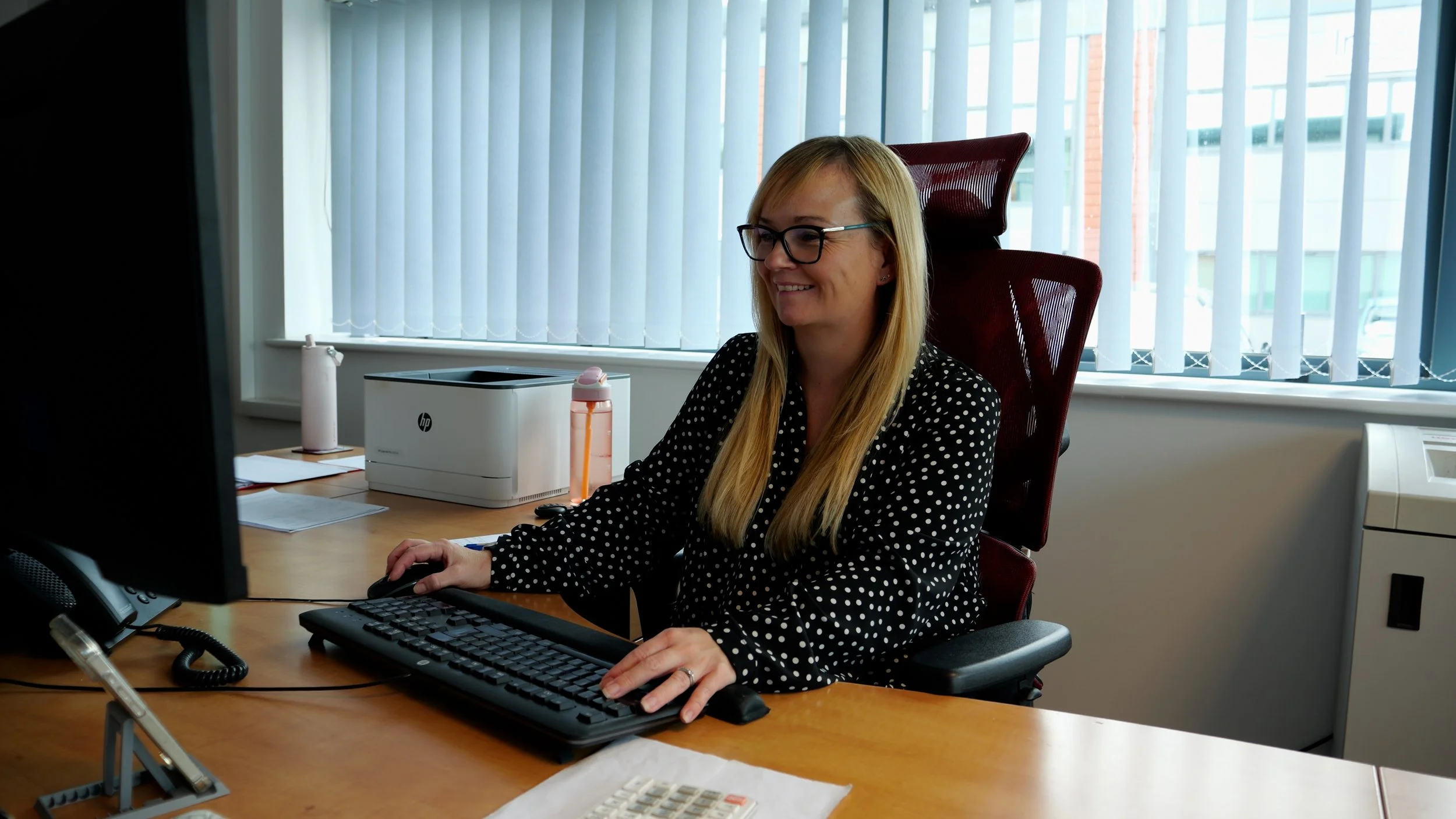 A woman with blonde hair and glasses working at a desk in an office, smiling at her computer monitor. She is wearing a black and white polka dot blouse, and the office has large windows with vertical blinds.