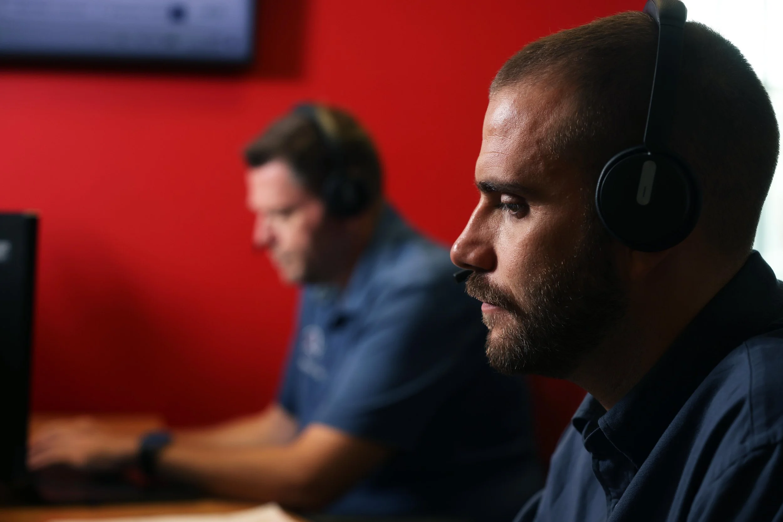 Two men wearing headsets working on computers in an office with a red wall background.