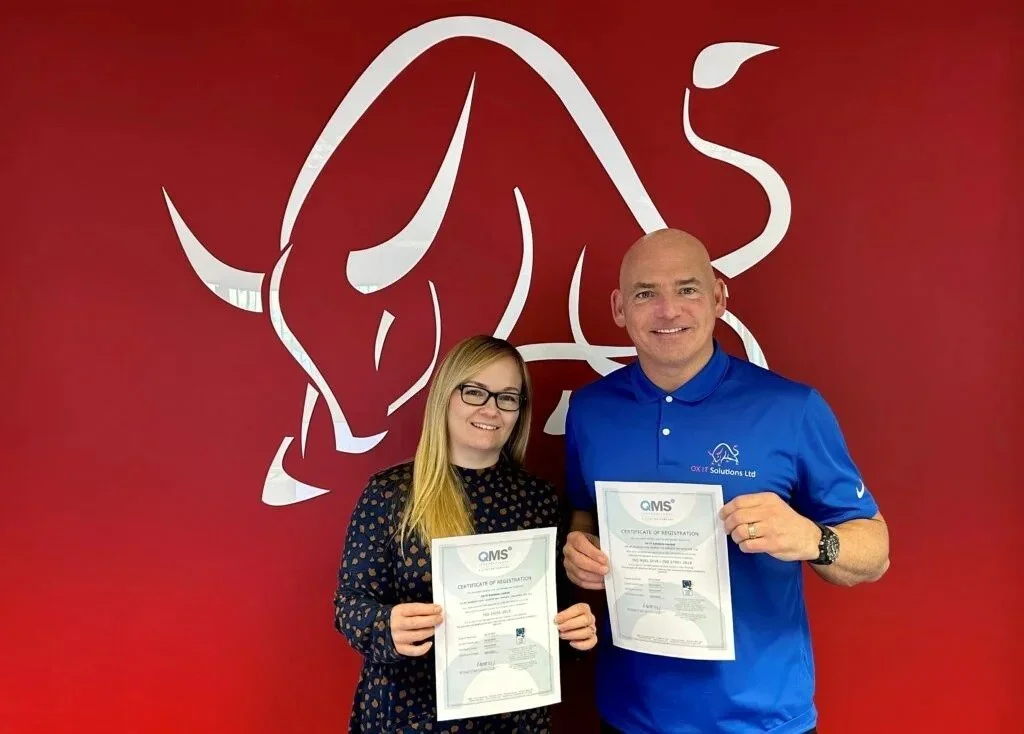 A woman and a man holding certification papers smiling in front of a red wall with a white bull logo.