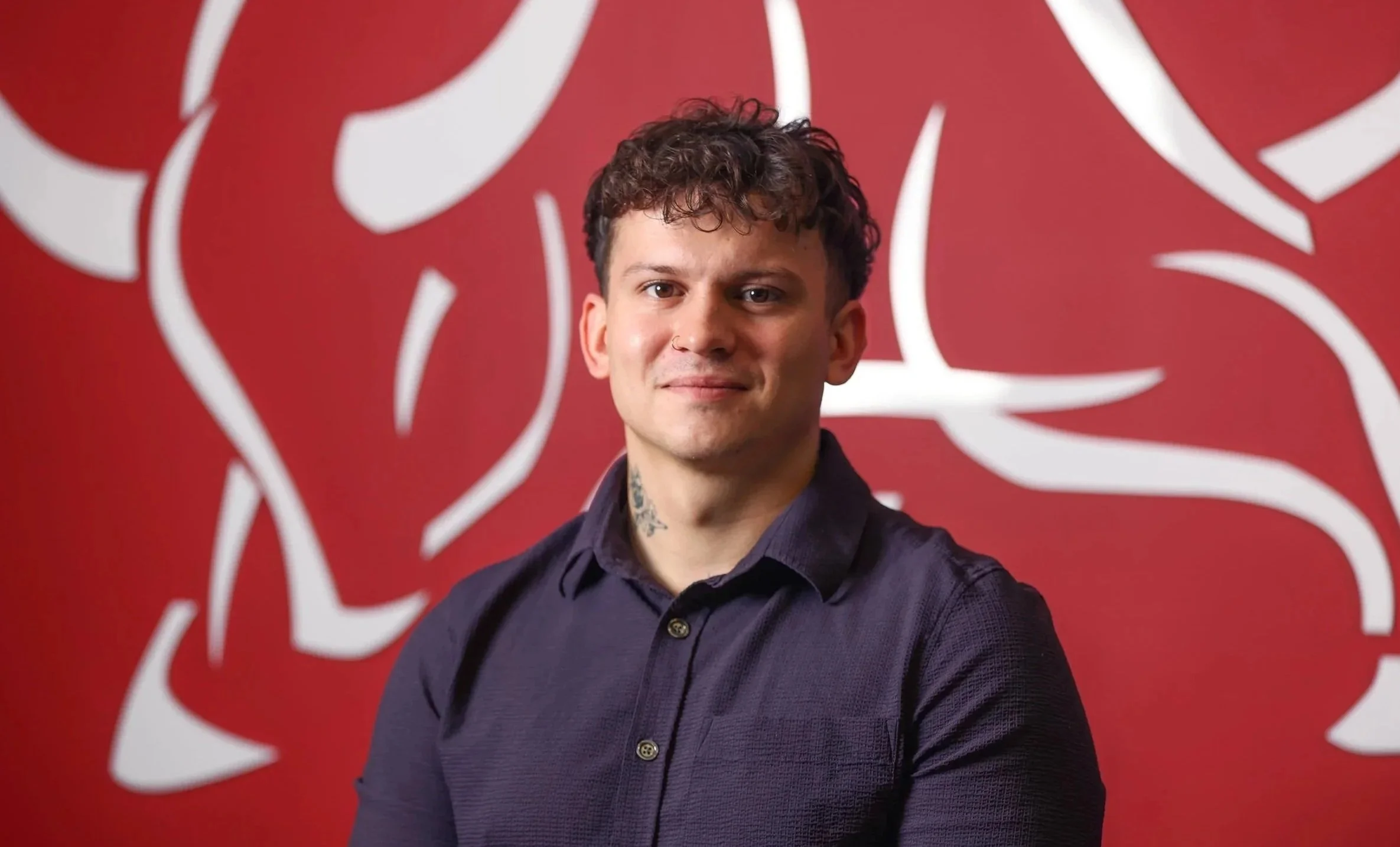 Portrait of a young man with curly hair and tattoos, wearing a dark button-up shirt, standing in front of a red background with white abstract designs.