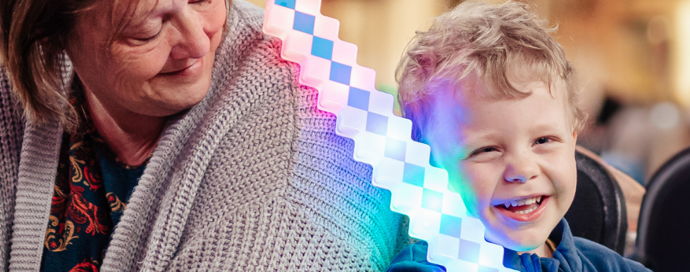 A smiling young boy with curly blonde hair and a woman with brown hair laughing together, sharing a colorful LED light stick.
