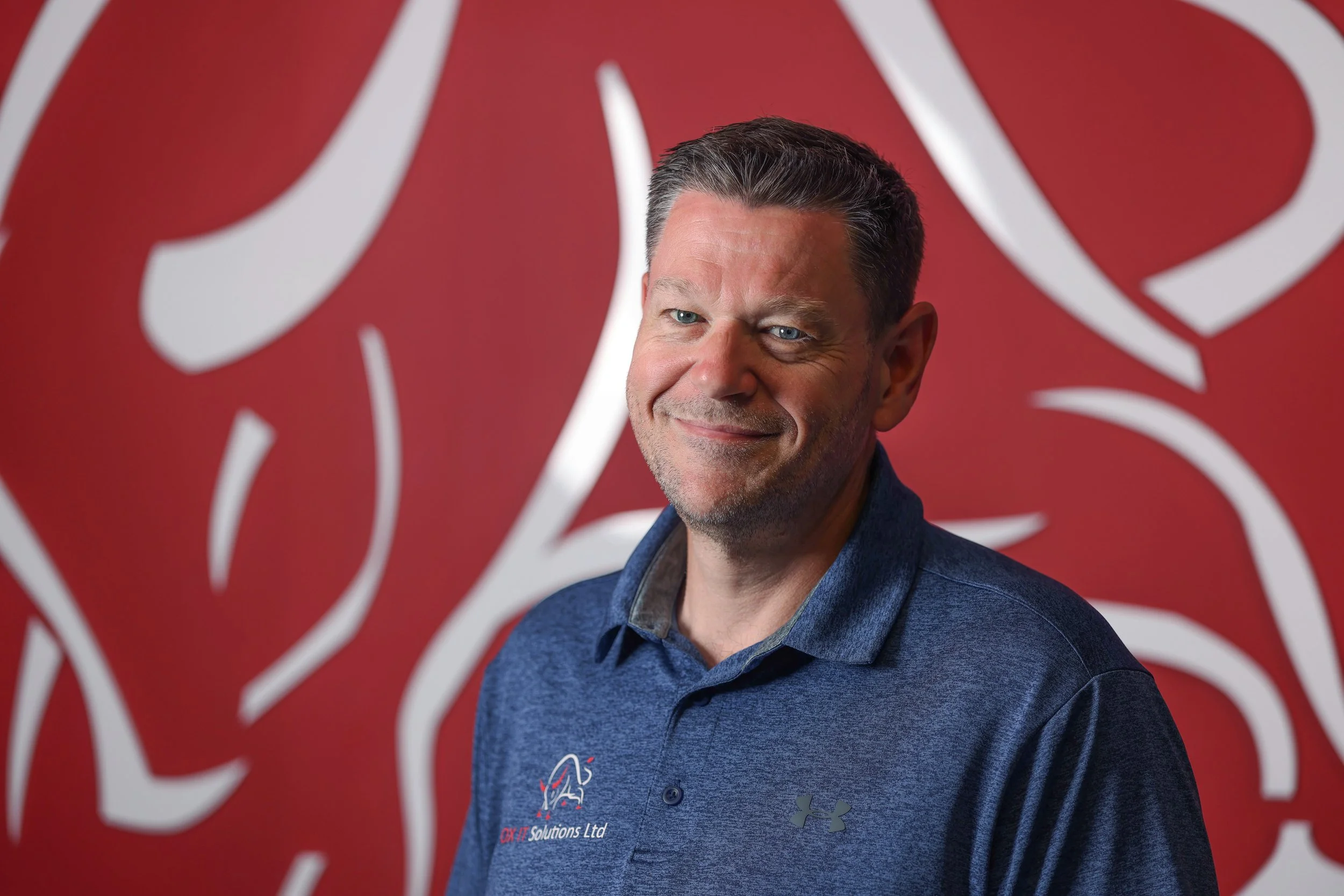 A man with short dark hair and blue eyes smiling and wearing a dark blue sport coat and shirt, standing in front of a red background with white abstract lines.