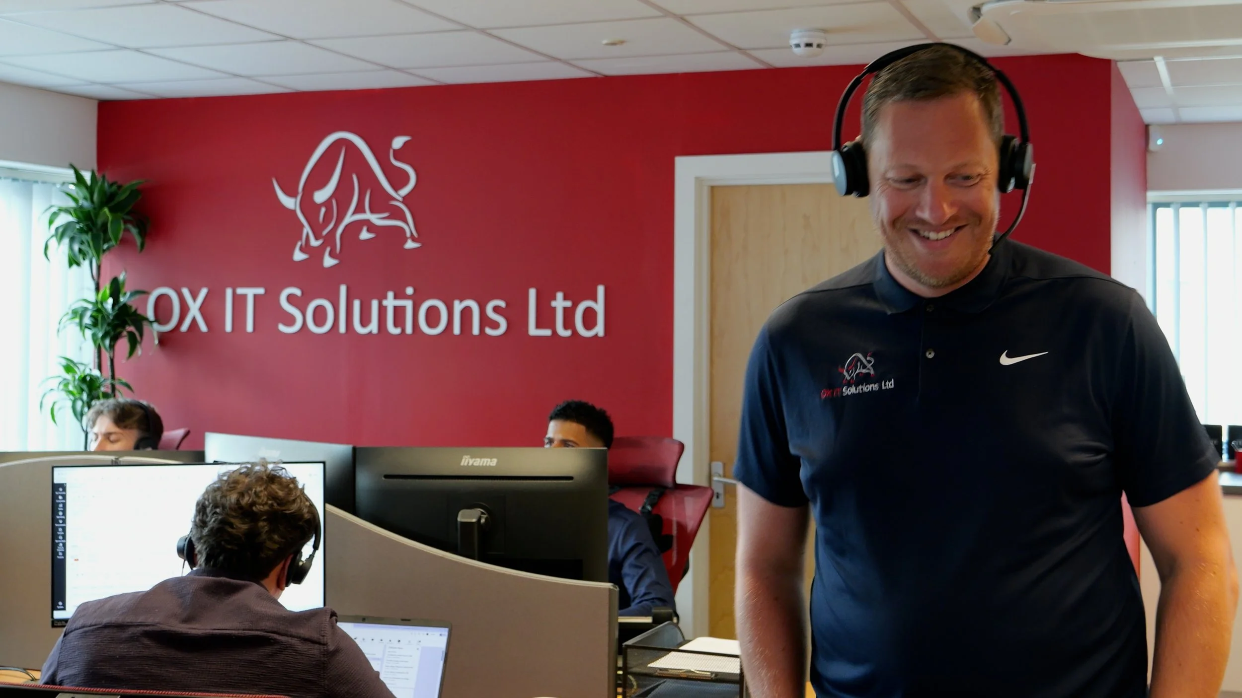 An office environment with multiple computer workstations and people working at their desks. A red wall in the background displays the logo and name 'OIX IT Solutions Ltd' with a stylized bull graphic. A man wearing a black Nike polo shirt with the company's logo and headset is smiling, walking through the office.