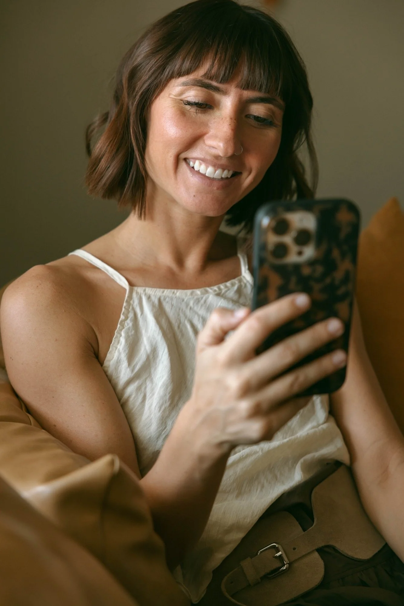A woman with shoulder-length brown hair and freckles is smiling and looking at her phone, wearing a white sleeveless top.