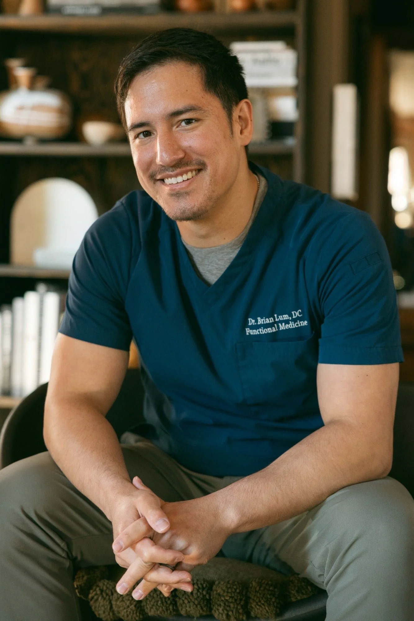 A smiling man with short dark hair and a goatee, wearing a navy blue medical scrubs shirt with white embroidery, sitting with his arms crossed on his lap, in an indoor setting with wooden shelves and various decorative objects in the background.