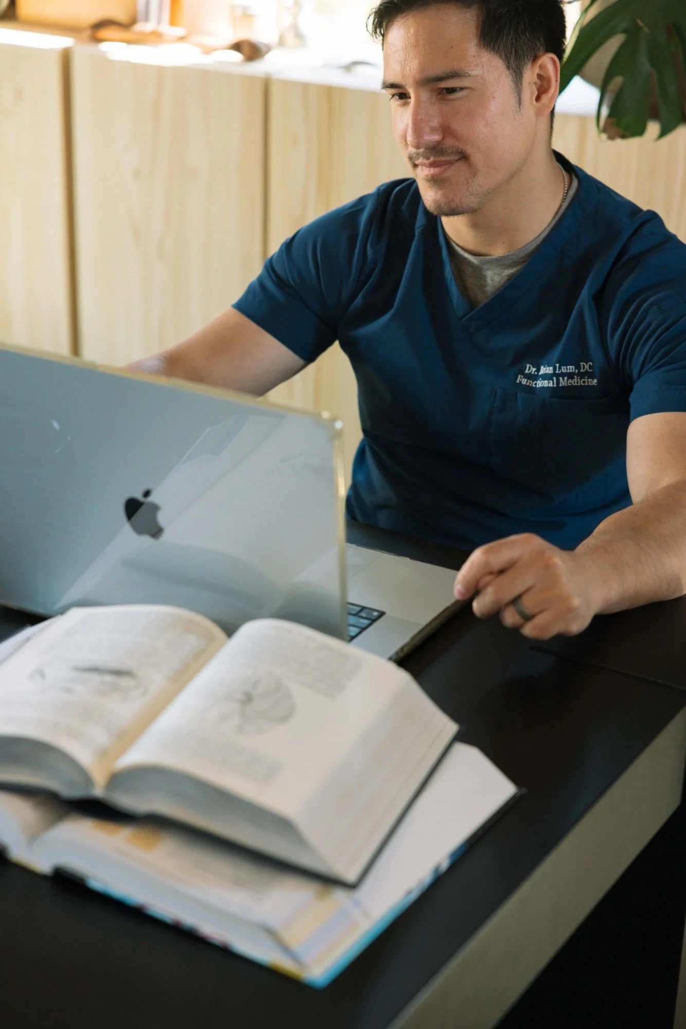 A medical professional wearing a navy scrub top with a name tag that reads 'Dr. Alan Lum, DC Functional Medicine' is sitting at a desk with an open laptop, looking at a book or documents, with a background of a wooden furniture and a potted plant.