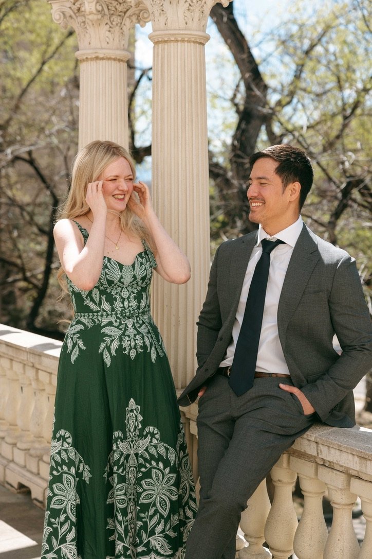 A woman in a green dress with white floral patterns laughs with a man in a gray suit and black tie, standing outdoors near classic white columns and a balustrade with trees in the background.