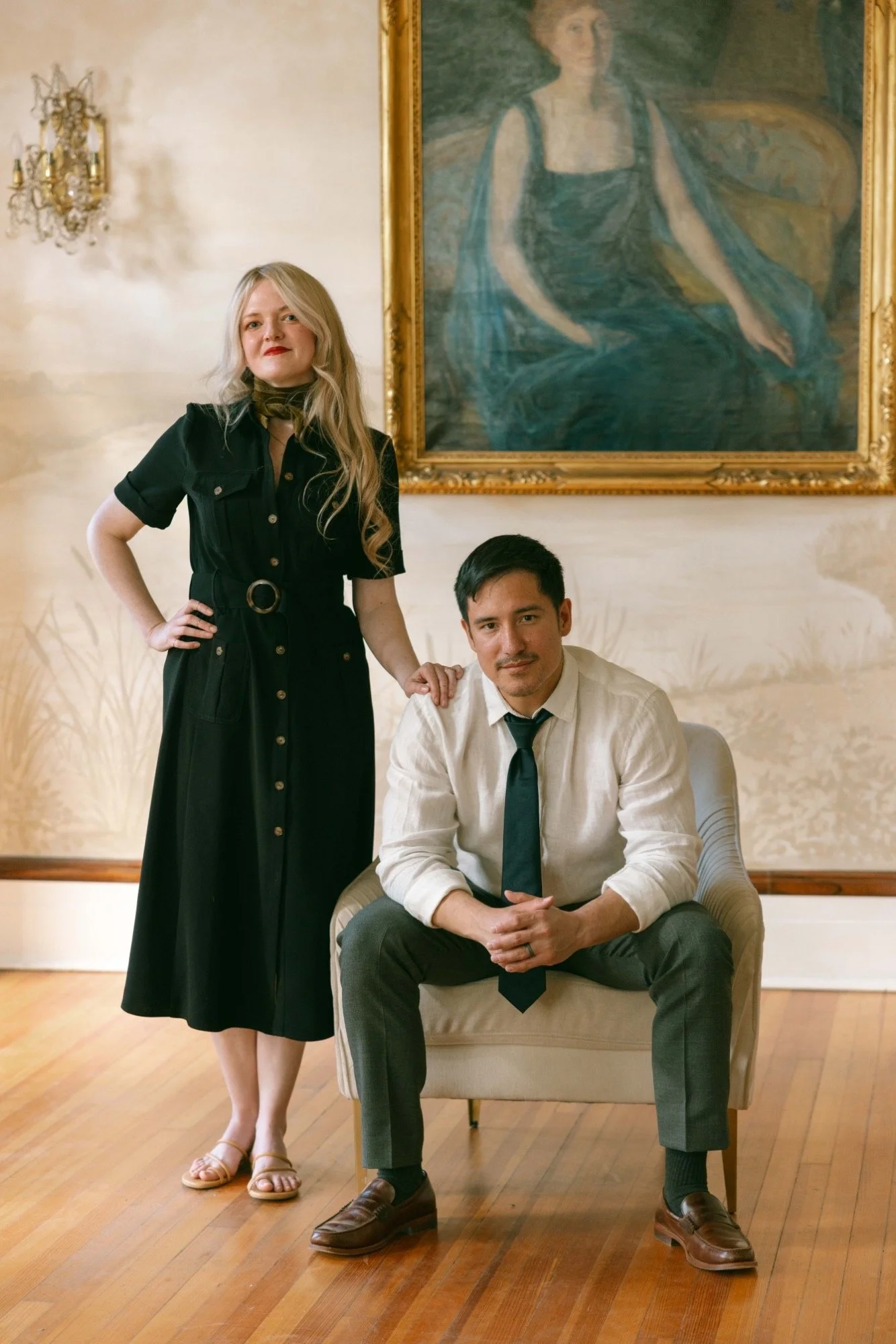A woman with long blonde hair in a black dress standing with her hand on her hip next to a seated man with dark hair in a white shirt and black tie. They are in a room with wooden floors, a large painting of a woman in a black dress hanging on the wall, and ornate chandeliers.