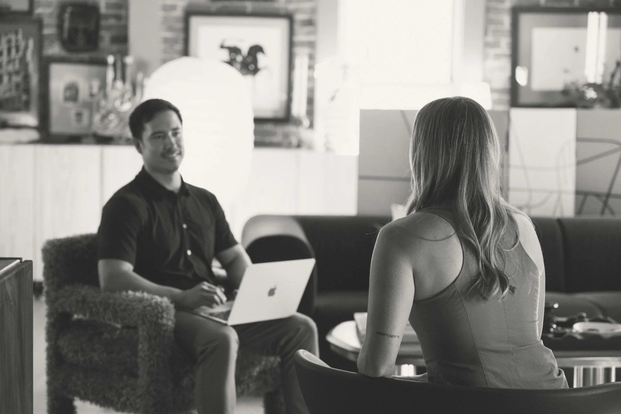 A woman sitting and facing a man in a casual office setting, with the man smiling and the woman turned away, both engaged in conversation, with computer screens and home decor in the background.