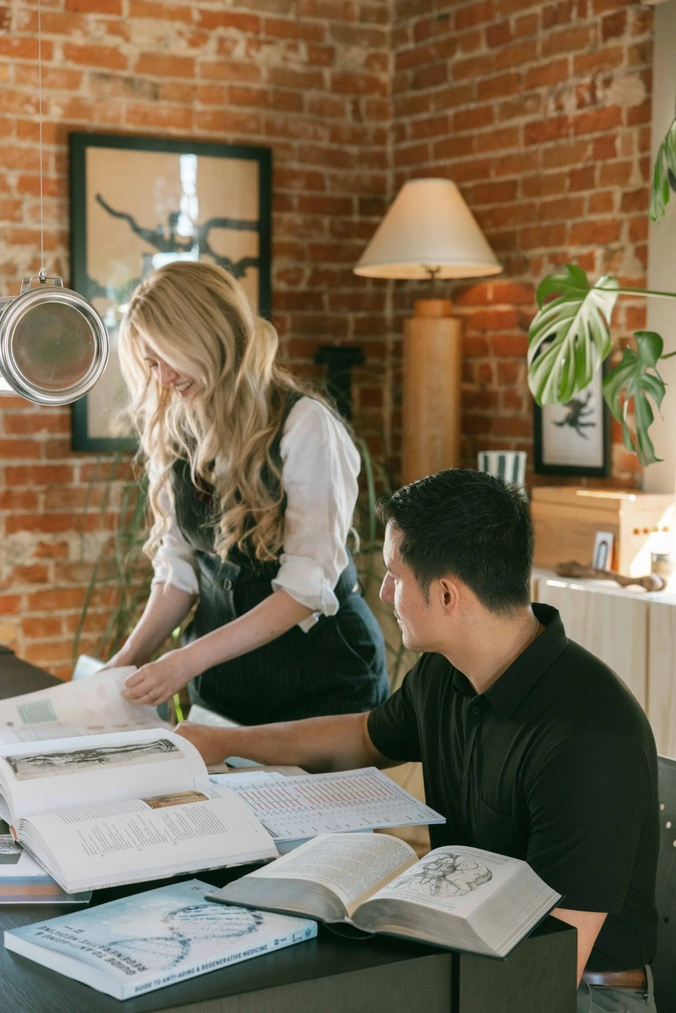 Two people studying at a table in a room with brick walls, books, and scientific diagrams open on the table. A woman with long blonde wavy hair is standing and smiling, while a man with short black hair is sitting and looking at the books.