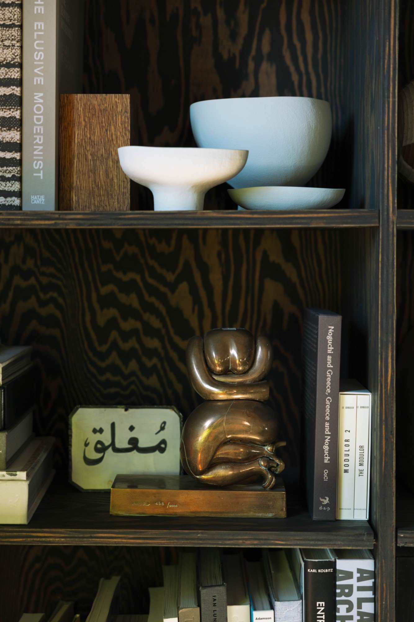 A wooden bookshelf with decorative objects and books, including a brass sculpture resembling a clenched fist, ceramic bowls, and various hardcover and paperback books.
