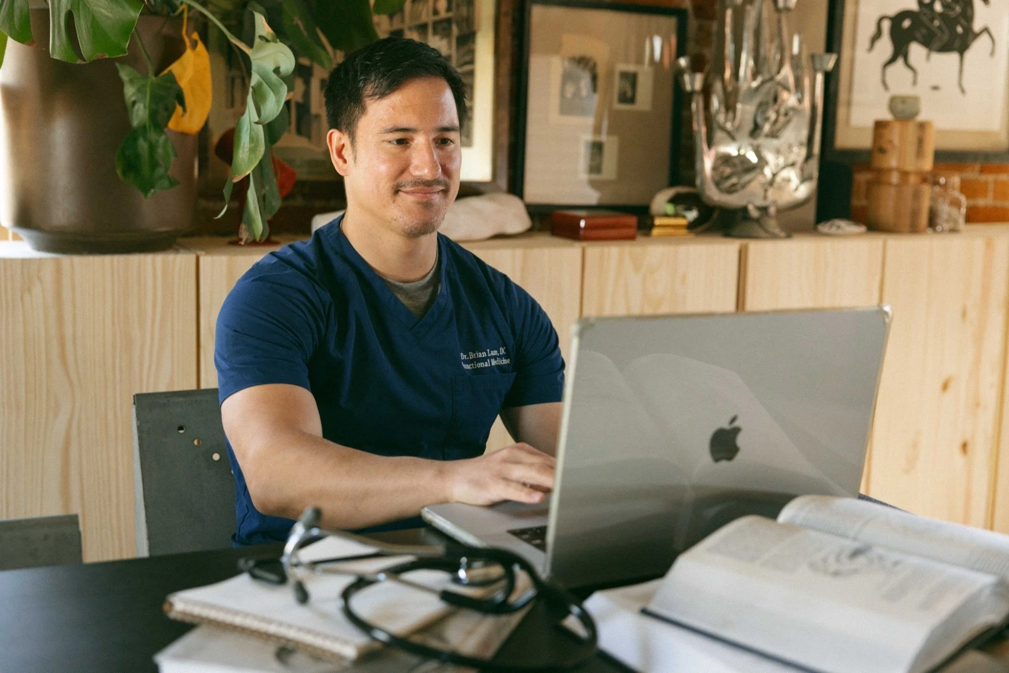 A man in a blue shirt, sitting at a desk with a silver MacBook laptop in front. Behind him is a wooden shelf with various decorative items, framed pictures, and artwork on the wall.