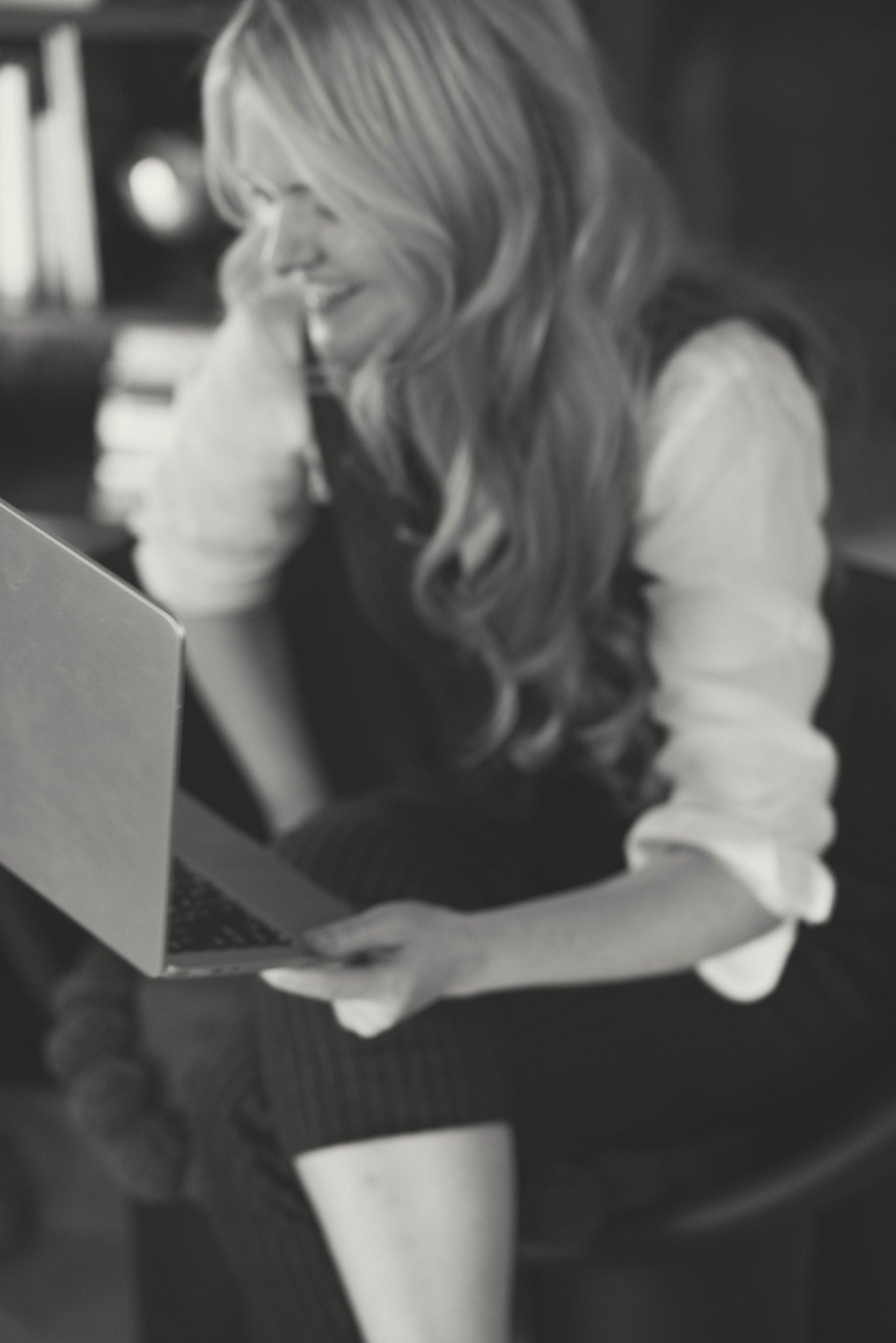 A woman with long, wavy hair smiling and looking at a laptop or tablet in front of her, with a blurred background.