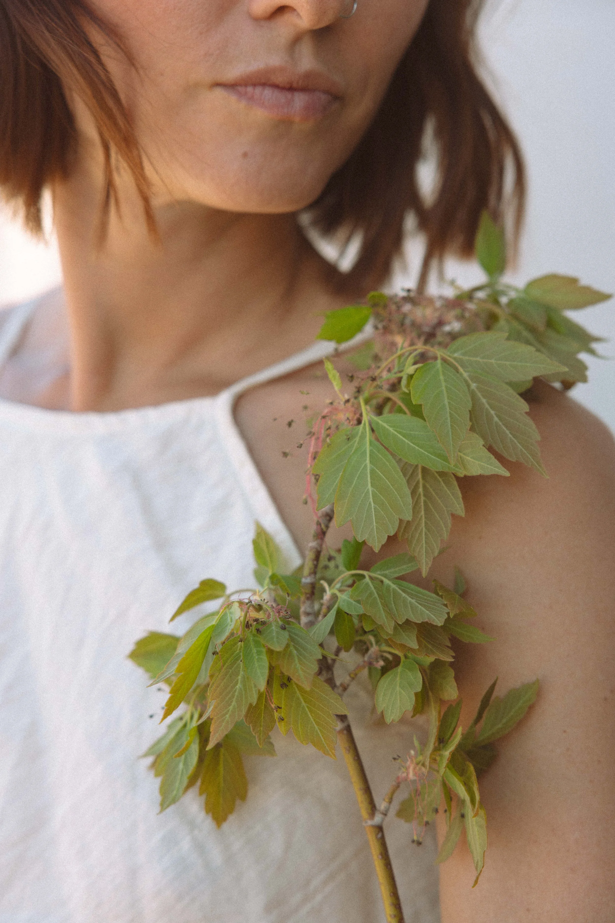 A close-up of a woman with reddish-brown hair, wearing a white sleeveless top, holding a branch with green leaves and pink flowers.