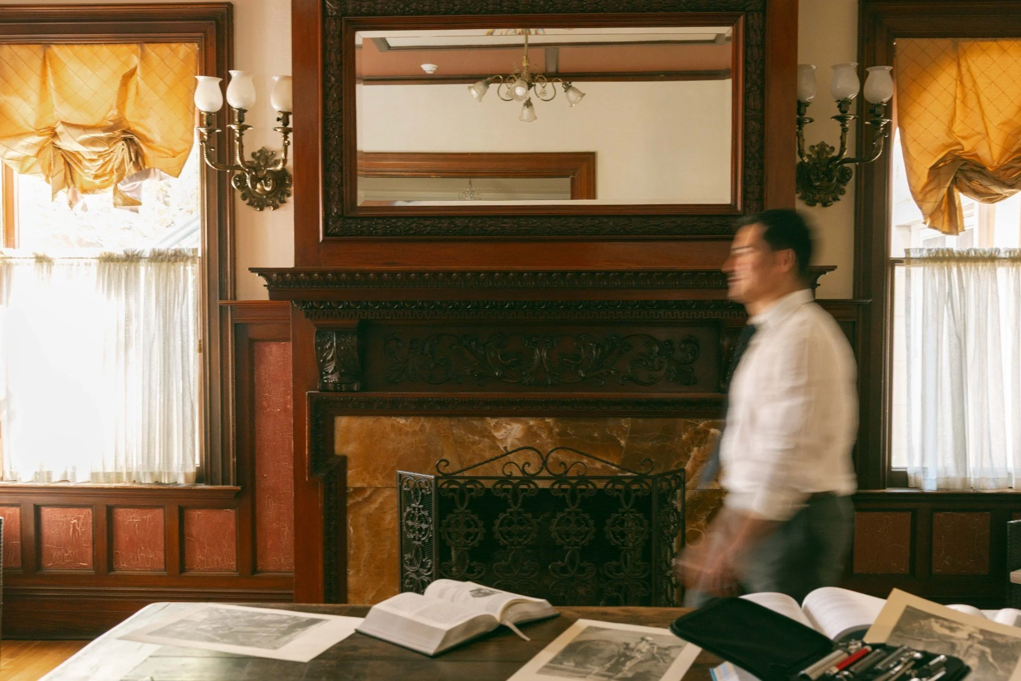 Interior of a room with two windows covered with yellow curtains, two ornate wall-mounted lamps, a large mirror, and a fireplace with a decorative metal grate. A man in a white shirt walks past, and a table in the foreground has open books and papers.