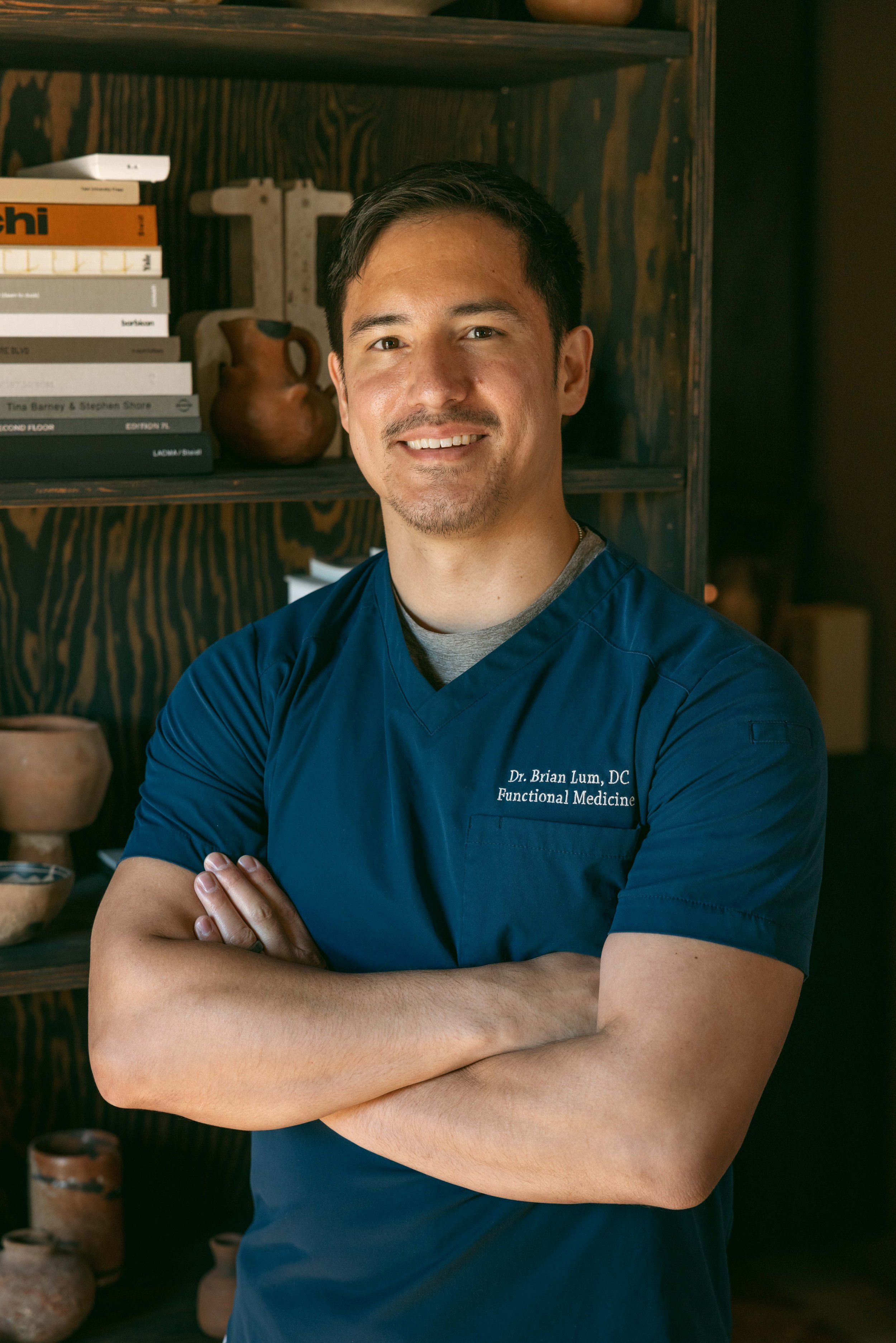 A man with dark hair and light skin, wearing a blue medical scrub top, standing with his arms crossed in front of a wooden bookshelf filled with books and pottery. He is smiling at the camera.
