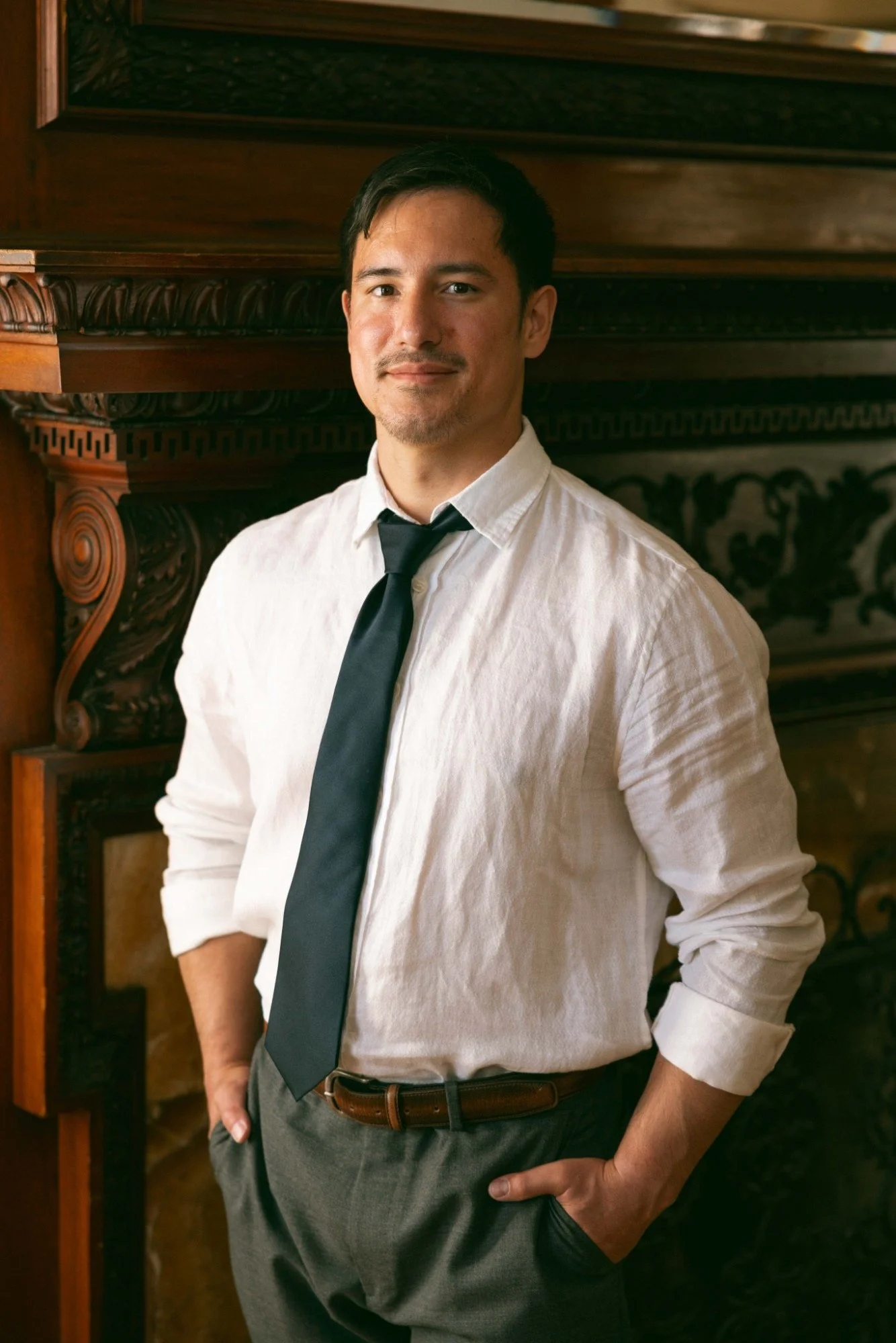 A young man in a white shirt, black tie, and gray pants, standing indoors in front of a wooden ornate fireplace.