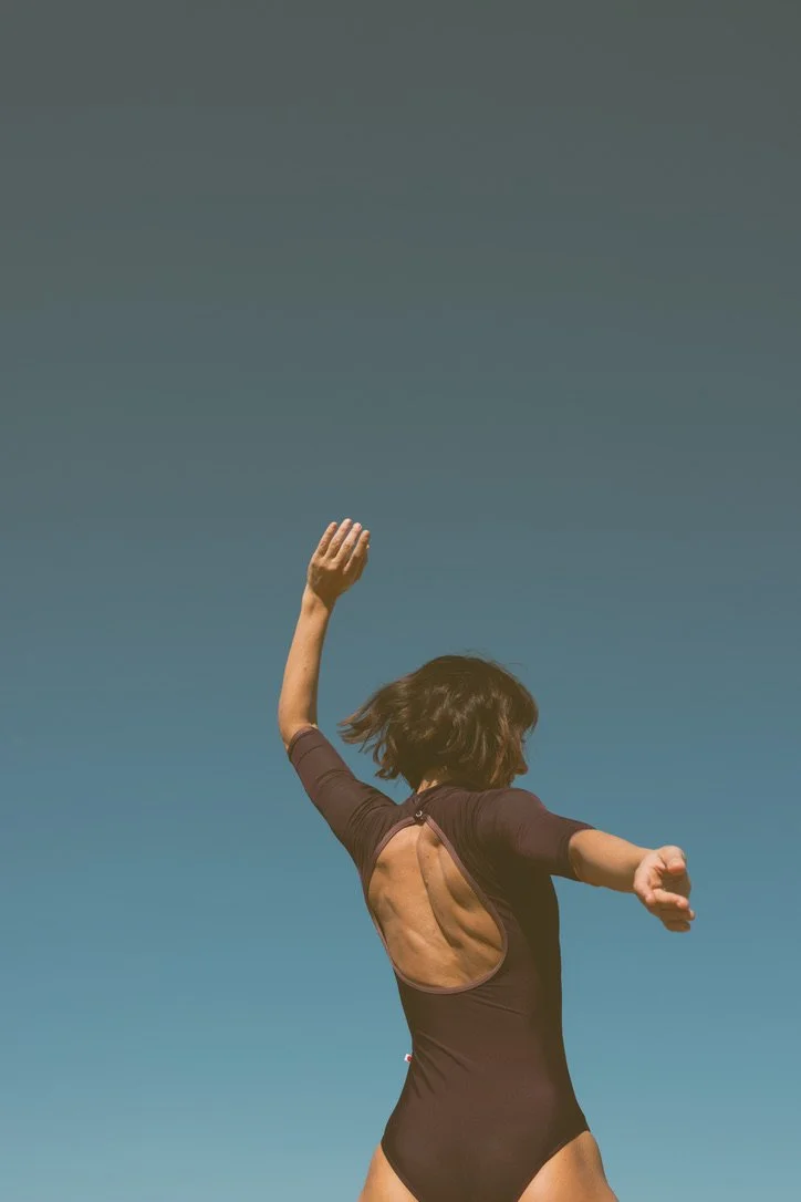 A woman in a black swimsuit performing a dance or pose outdoors against a clear blue sky.