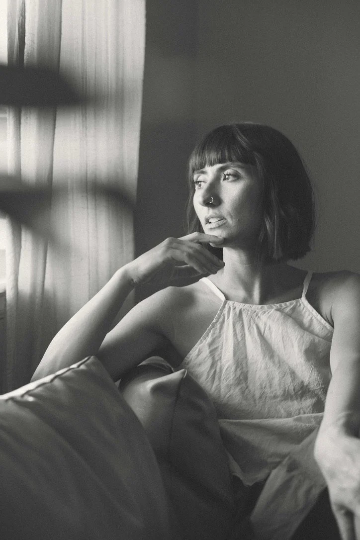 Black and white photo of a woman sitting on a sofa, gazing out of a window with curtains, resting her chin on her hand, with a contemplative expression.