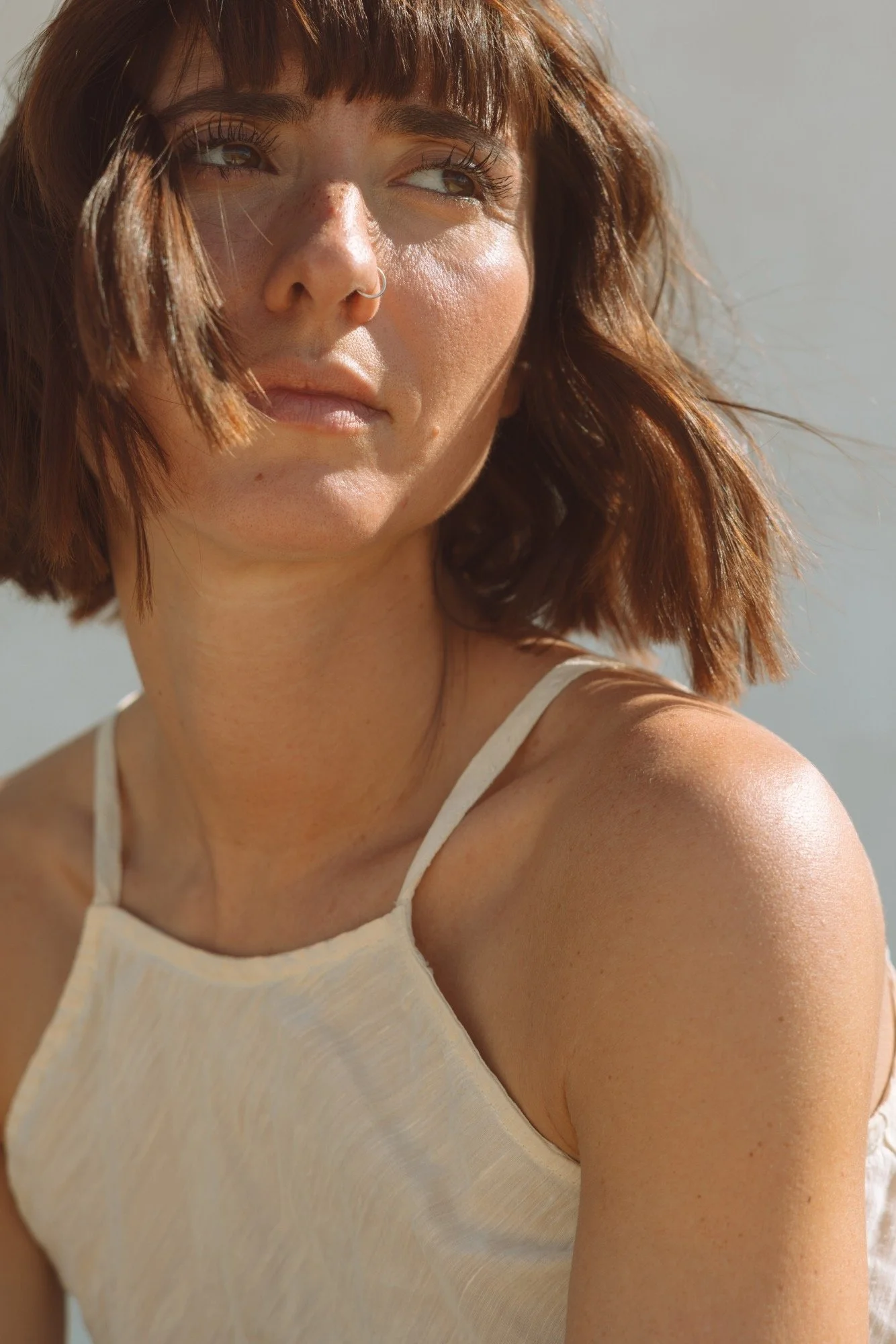 Close-up of a smiling woman with red hair, light skin, freckles, wearing a light-colored sleeveless top, outdoors in natural sunlight.