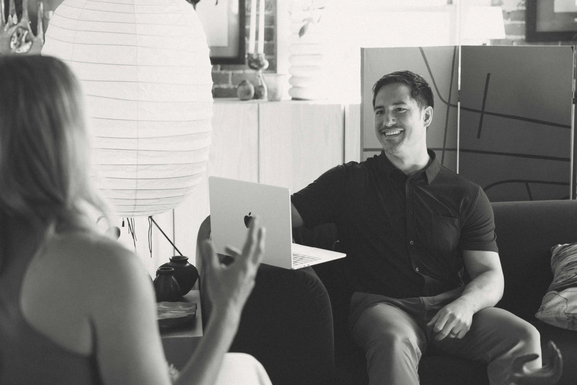 A man with short dark hair and a dark shirt smiling during a conversation with two women sitting at a table. One woman is holding a smartphone, and the other is partially visible in the foreground. The setting appears to be indoors with decorative lamps and a brick wall background.