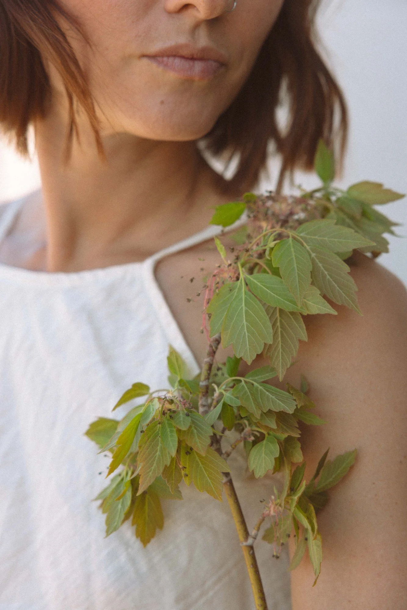 Close-up of a woman's face with shoulder-length brown hair, wearing a white sleeveless top, holding a branch with green leaves and pink flowers.