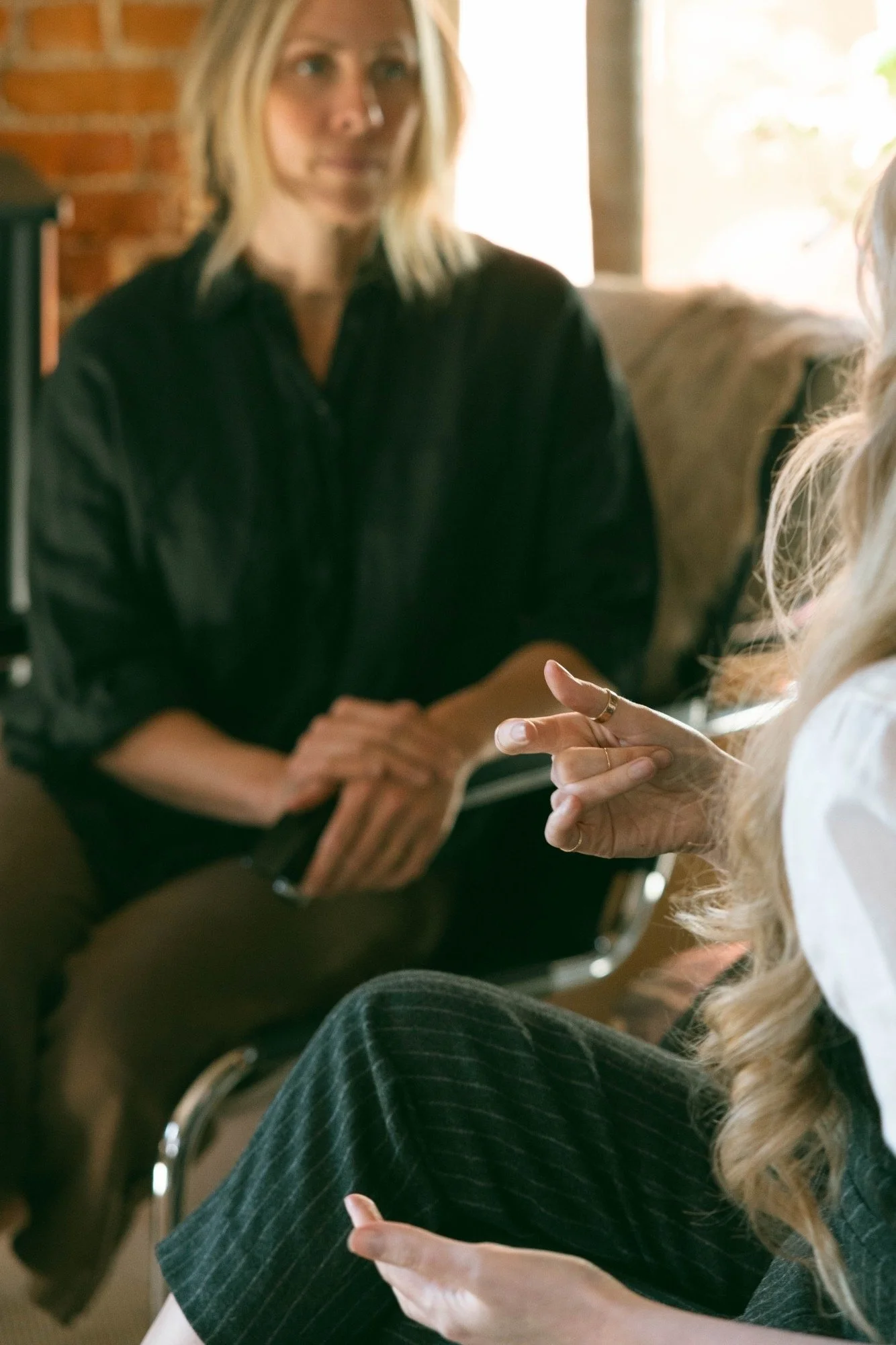 Two women sitting indoors, engaged in conversation. One woman has long blonde hair and is wearing striped pants, while the other woman has shoulder-length blonde hair and is wearing a black top. The background includes a brick wall and a window with sunlight coming through.