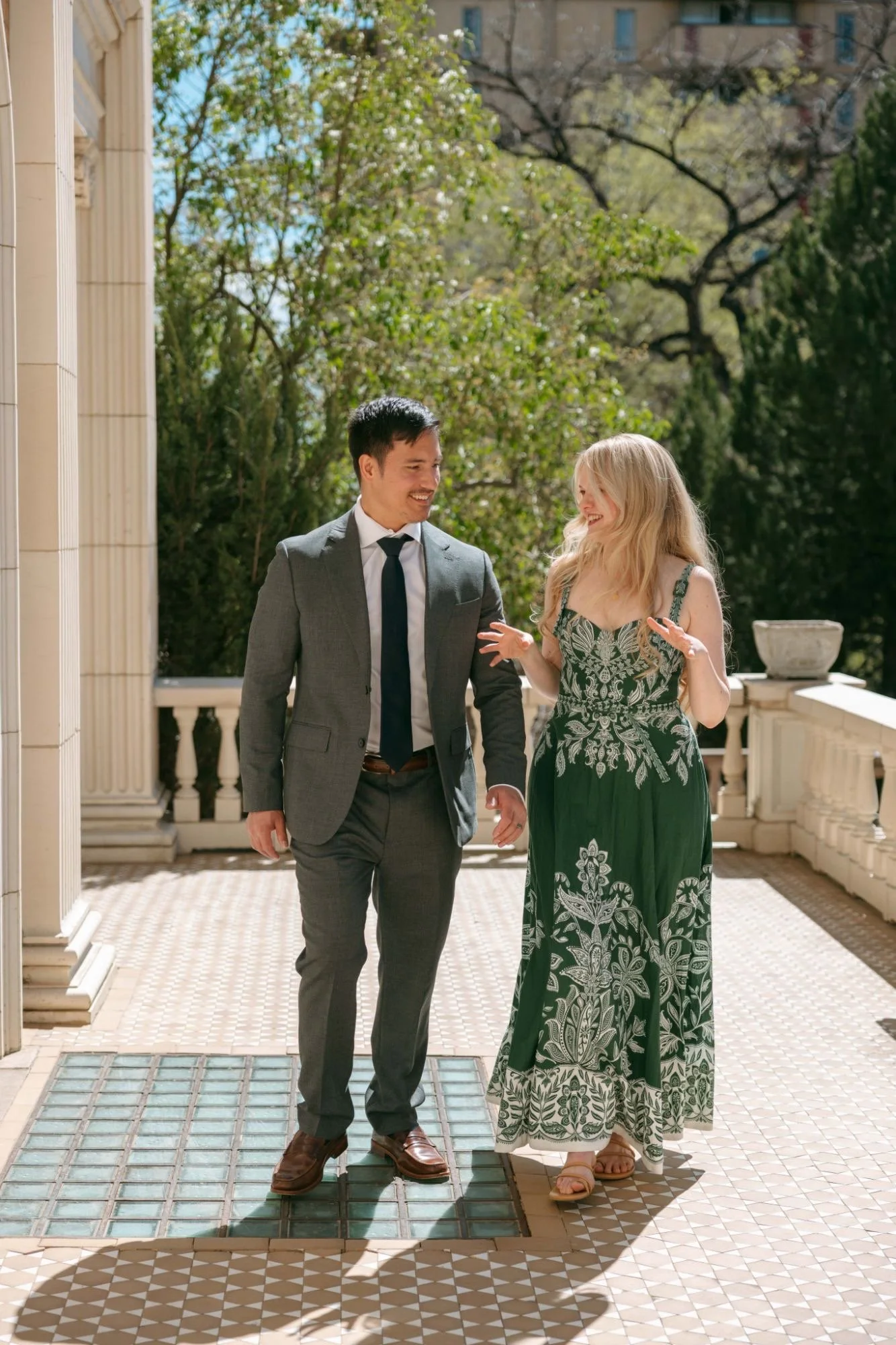 A young man in a grey suit and a young woman in a green dress are walking together outdoors on a sunny day, smiling and talking. They are on a balcony with columns and a patterned tile floor, surrounded by green trees.