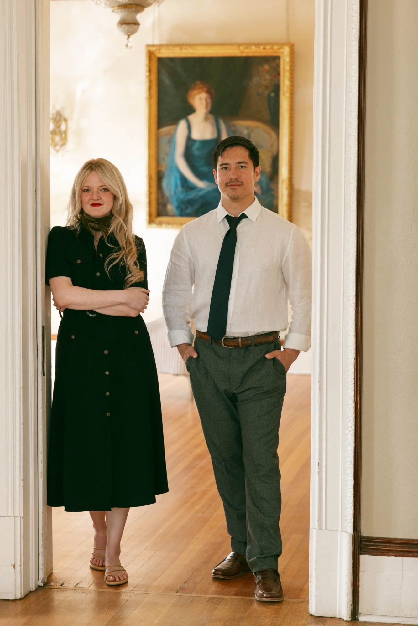 Two people, a woman with long blonde hair in a black dress with a scarf and a man with dark hair in a white shirt and black tie, stand inside a doorway with a painting of a woman in a blue dress hanging on the wall behind them.