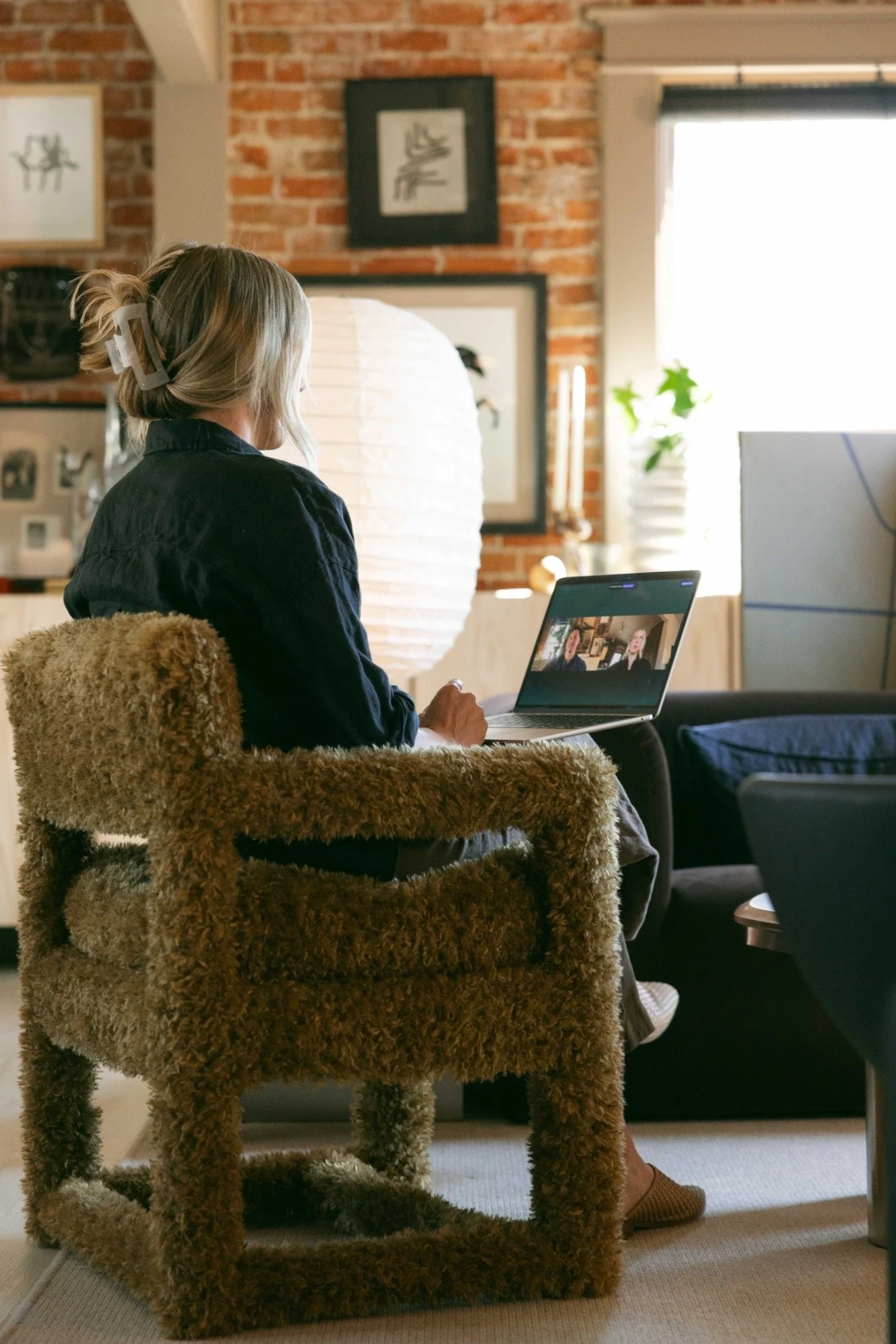 A woman with blonde hair, tied back with a large hair clip, is sitting in a cozy living room, looking at a laptop screen showing a video call. She is seated on a fluffy, brown dog-shaped chair. The room features a brick wall, framed pictures, a white paper lantern light, a window with a plant, and various decorative items.