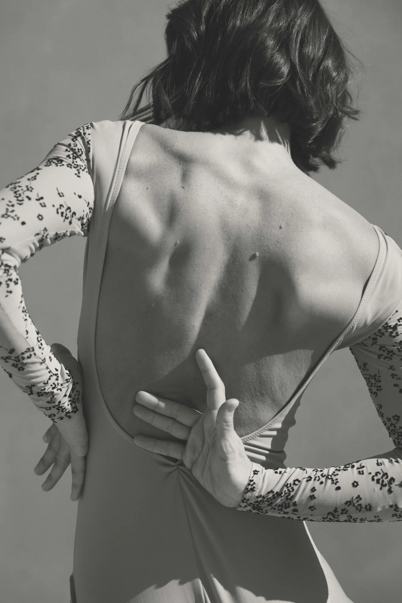 Black-and-white photo of a woman with short, wavy hair, wearing a long-sleeved top with leopard print on the sleeves. She is facing away, showing her back, with her right arm bent and hand resting on her back, and her left arm crossed and resting on her waist. A poised hand lower down is making a peace sign.
