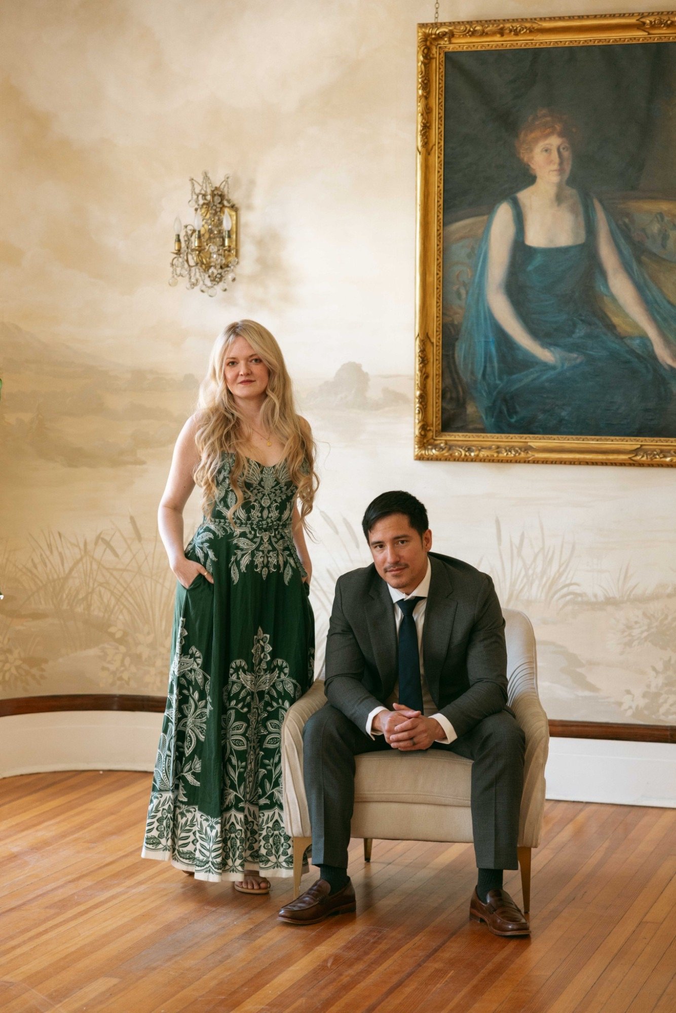 A woman in a green dress with white floral patterns standing next to a seated man in a suit. They are in a room with wooden floors, a beige armchair, a large painting of a woman in a blue dress with a gold frame, and a decorative wall sconce.