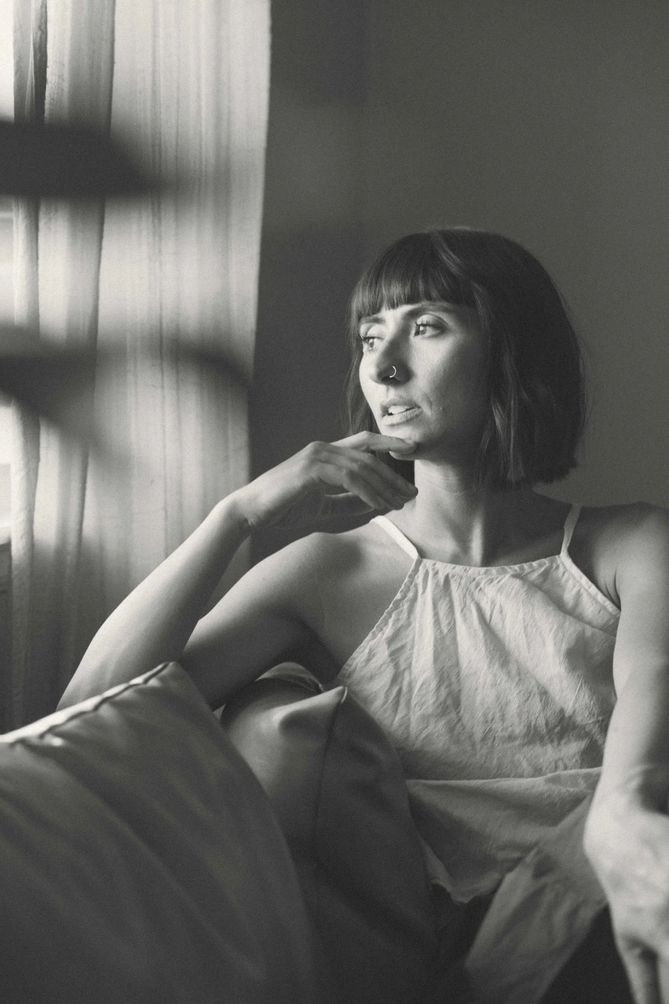 A black and white photo of a woman with dark hair and bangs, sitting by a window with vertical blinds. She has a nose piercing and is wearing a sleeveless top. She is resting her chin on her hand, looking thoughtfully out the window.