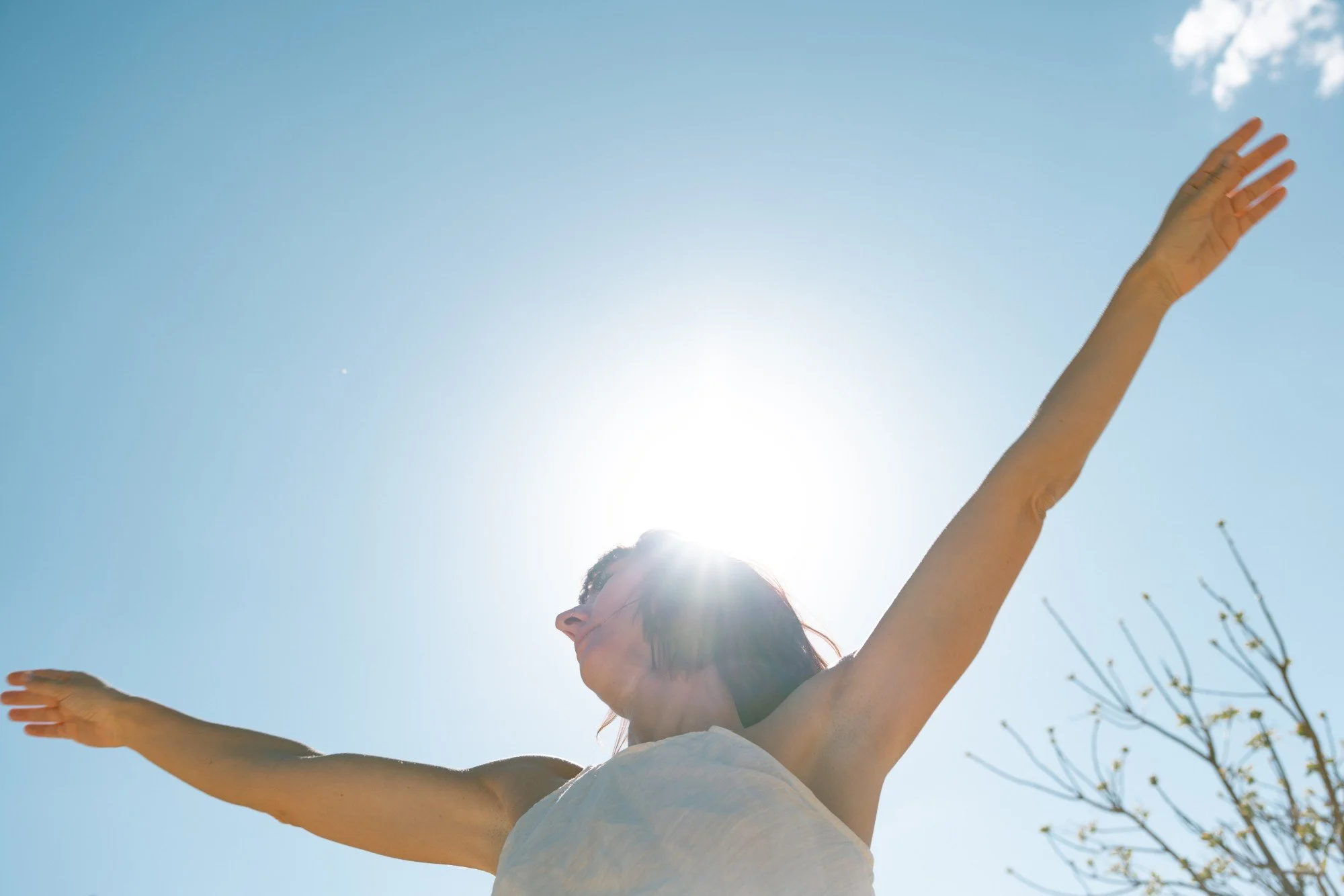 A woman with arms outstretched towards a clear blue sky, backlit by the sun.