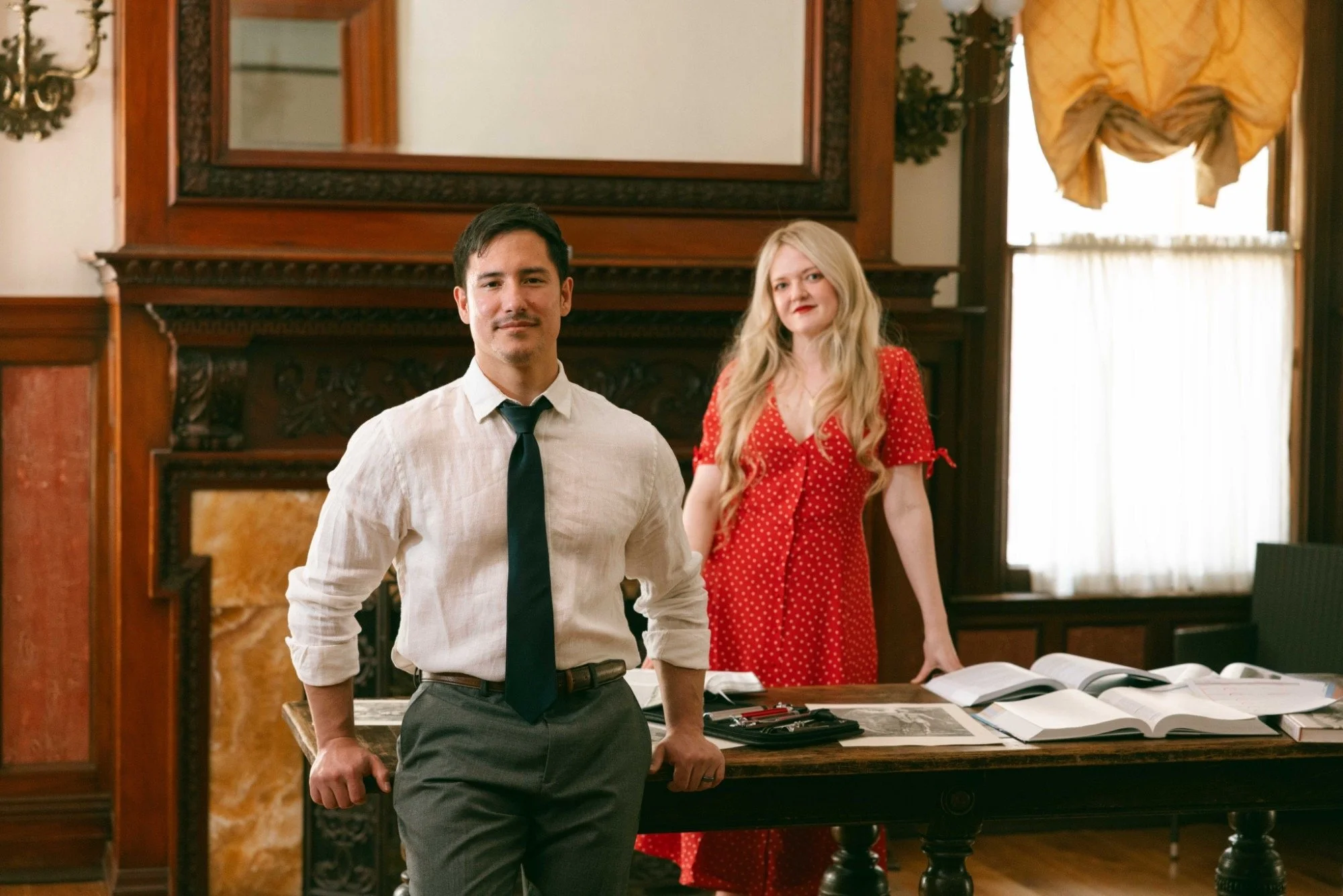 A man in a white shirt and dark tie standing in front of a woman in a red polka dot dress inside a room with ornate wooden paneling and a fireplace.