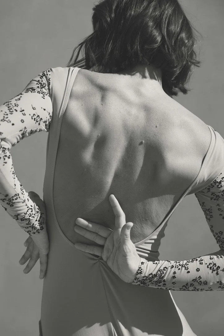 A woman with short, wavy hair posing with her back to the camera, wearing a long-sleeve top with lace details on the sleeves, exposing her back with her hands positioned on her waist and lower back.
