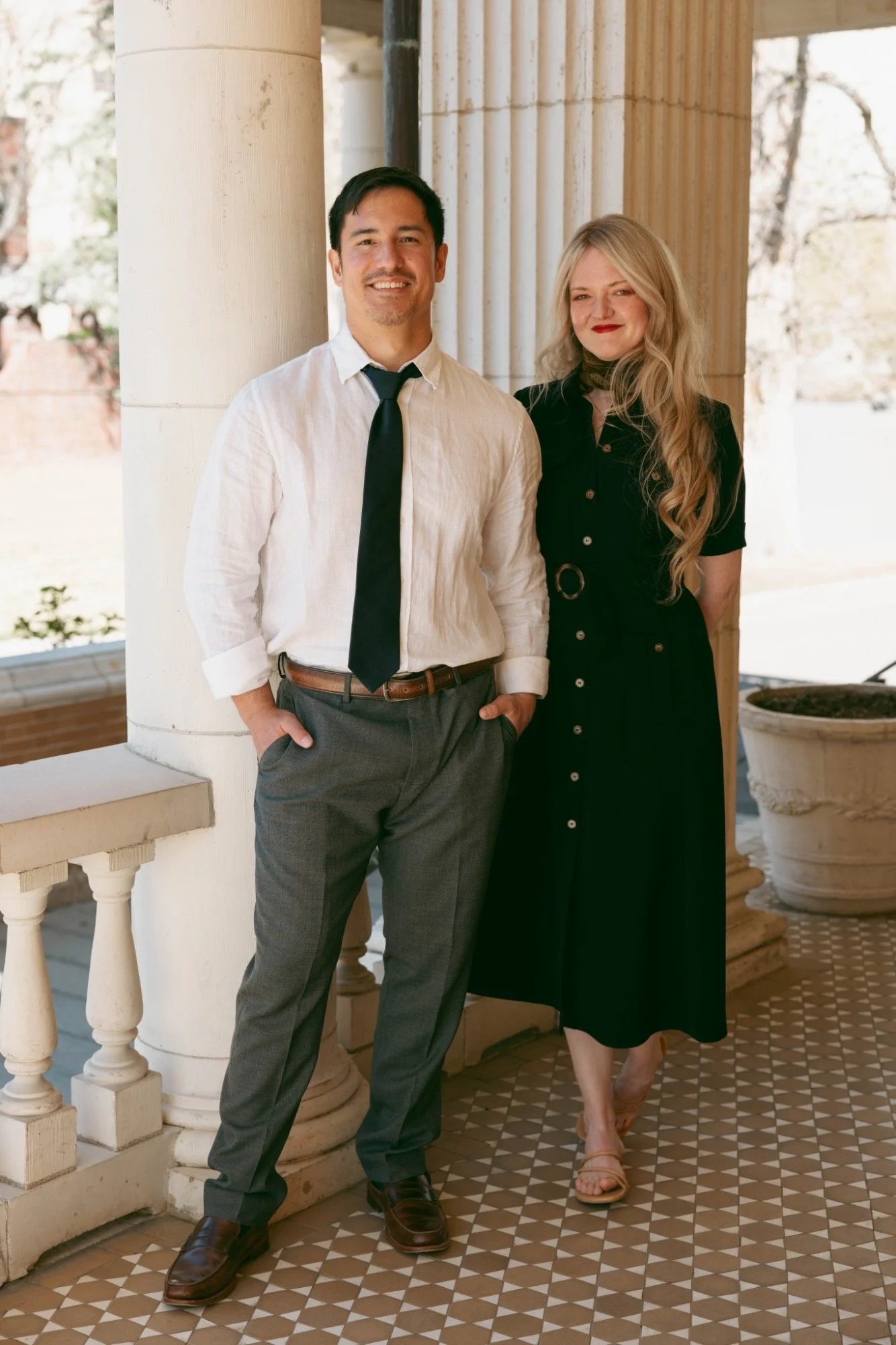 A man and a woman standing on a patterned tile floor, leaning against a white column with classical design, with trees visible in the background.