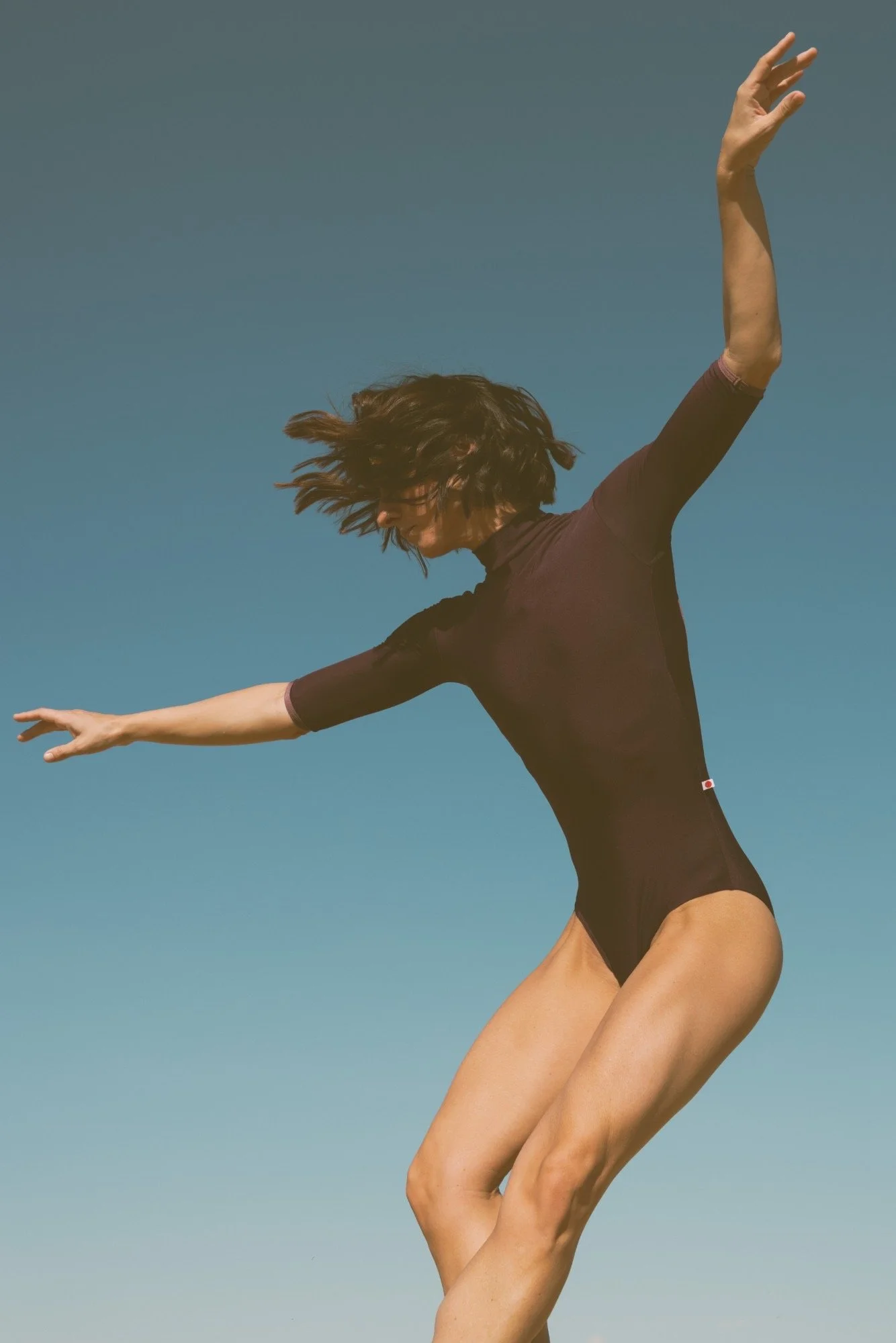 A person with short brown hair wearing a black leotard, seen mid-air with arms extended, against a clear blue sky.