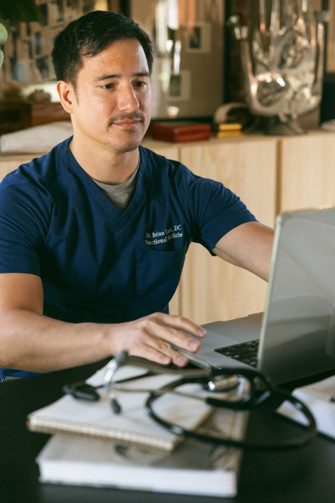 A man in scrubs working on a laptop at a desk with notebooks and a stethoscope, in a cozy, decorated room.