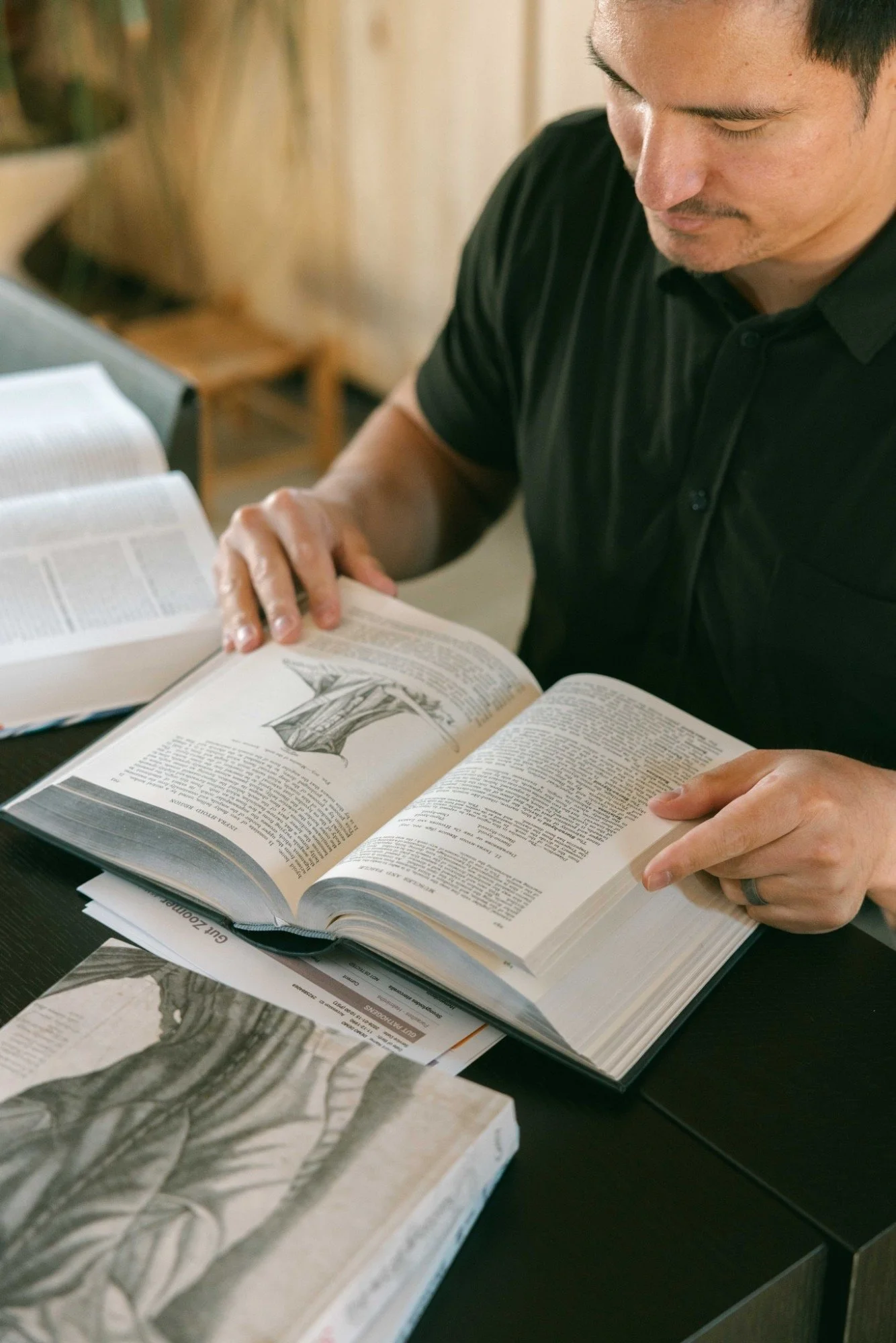A man reading a large book with detailed drawings, sitting at a table with additional books and papers around him, in a well-lit room.