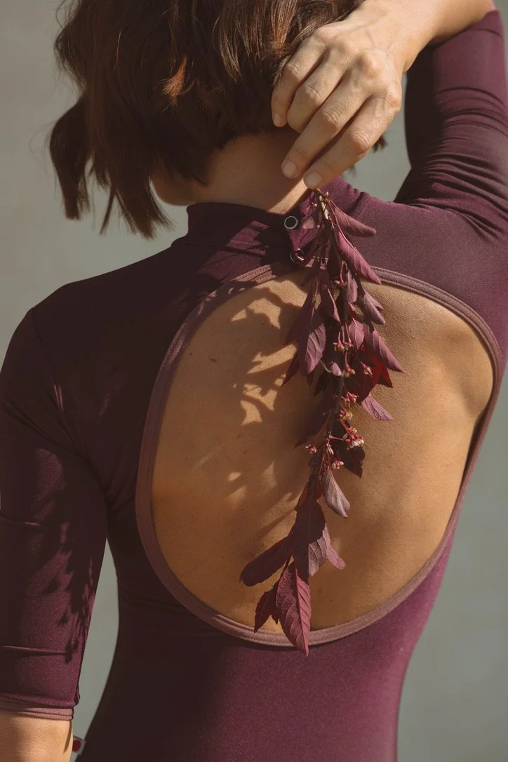 A woman with short brown hair adjusting her maroon athletic top with an open back, revealing decorative maroon leaves attached to the back of her shirt.