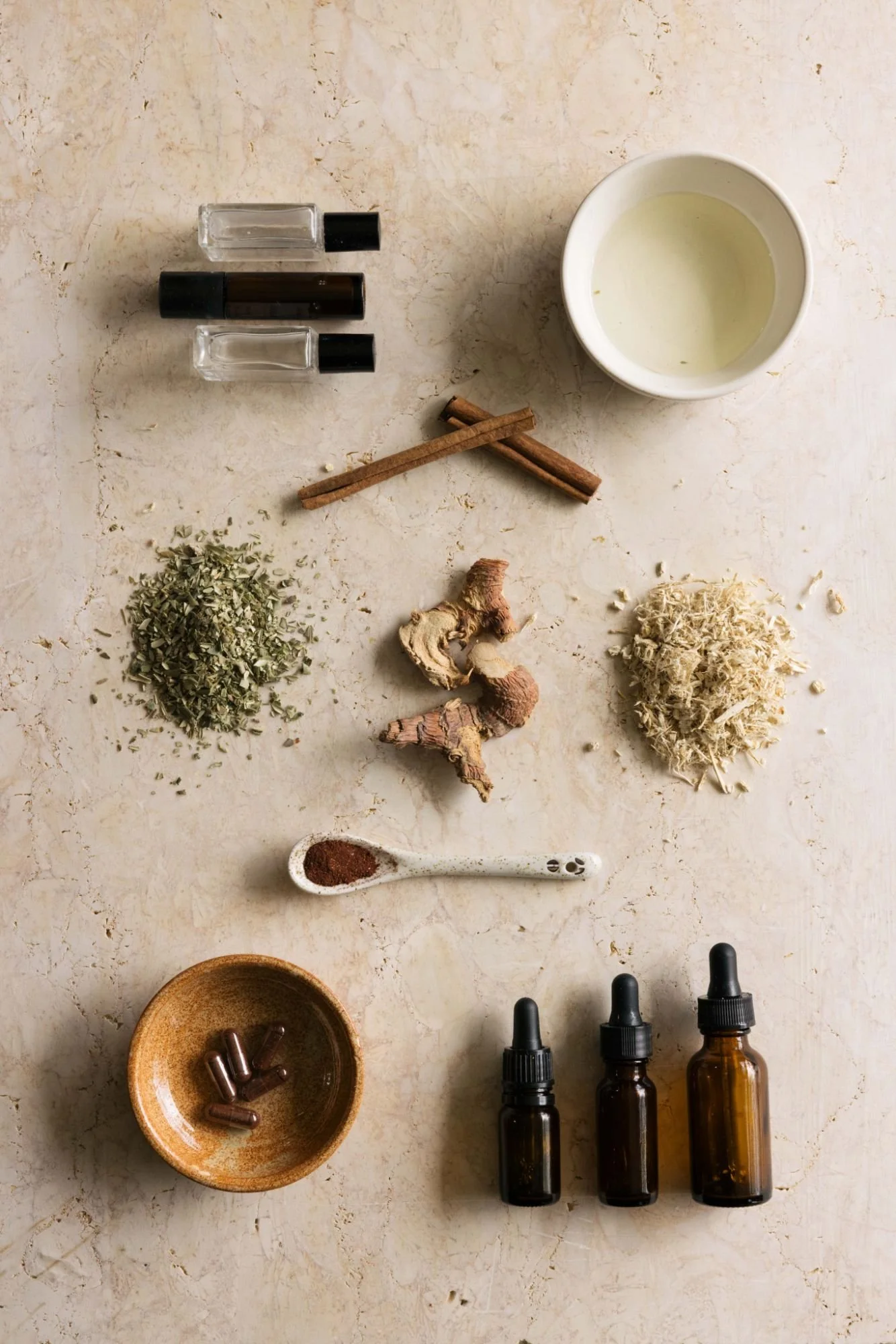 A flat lay of herbal ingredients, essential oils, and containers on a marble surface, including dried herbs, ginger, cinnamon sticks, dried flowers, a ceramic spoon with powder, and glass dropper bottles.