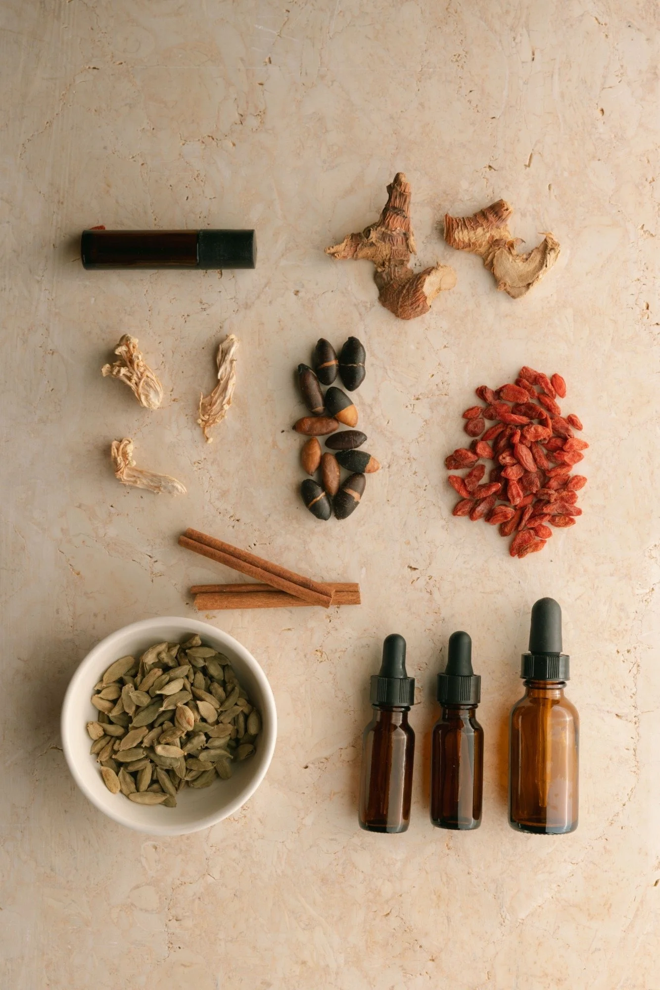 Various herbs and spices arranged on a marble surface, including dried ginger, goji berries, cardamom pods, cinnamon sticks, dried turmeric roots, black seeds, drops of essential oils, and dropper bottles.