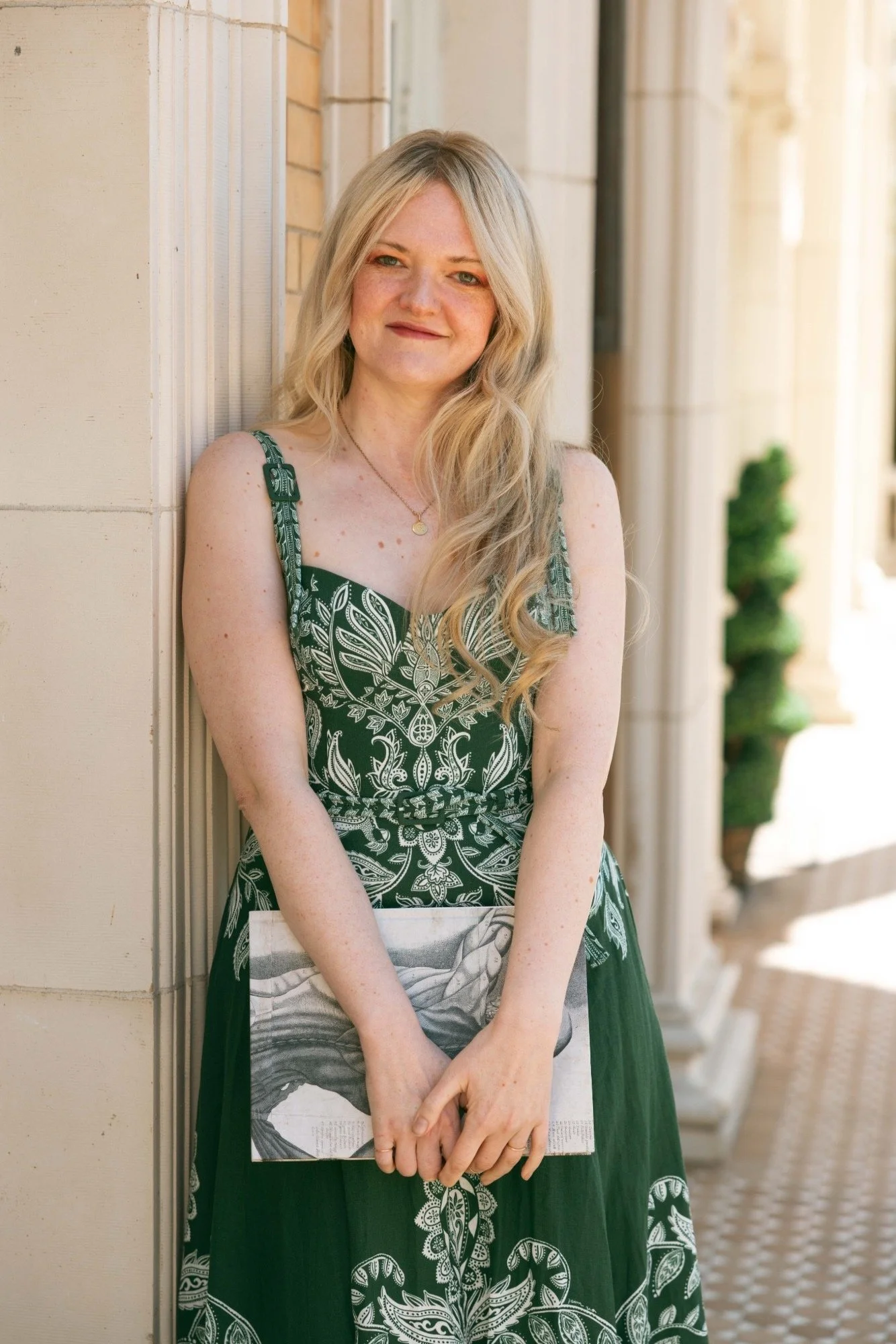 A woman with long blonde hair and freckles, wearing a green patterned dress and holding a newspaper, is standing outdoors against a beige wall, smiling at the camera.