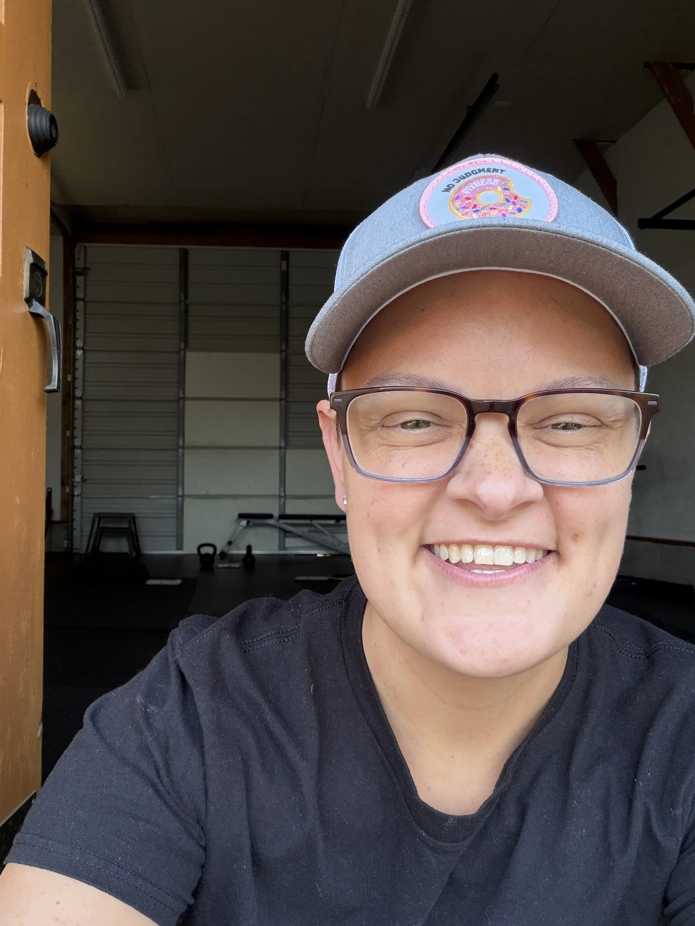 A woman smiling, wearing glasses and a light gray baseball cap with a donut patch on the front, inside a room with a garage door and workout equipment in the background.