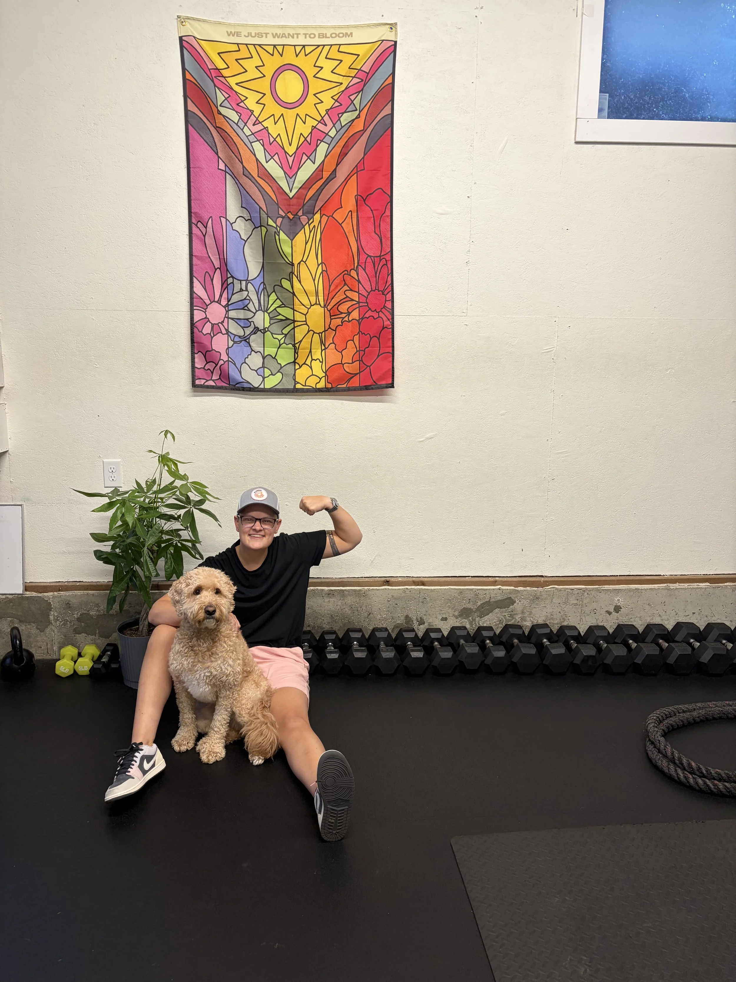 A person sitting on the floor in a gym, flexing their bicep and smiling, with a large fluffy dog sitting beside them. The person is wearing a black t-shirt, pink shorts, and a gray cap. There are dumbbells, a plant, and gym equipment in the background, along with a colorful abstract wall hanging.