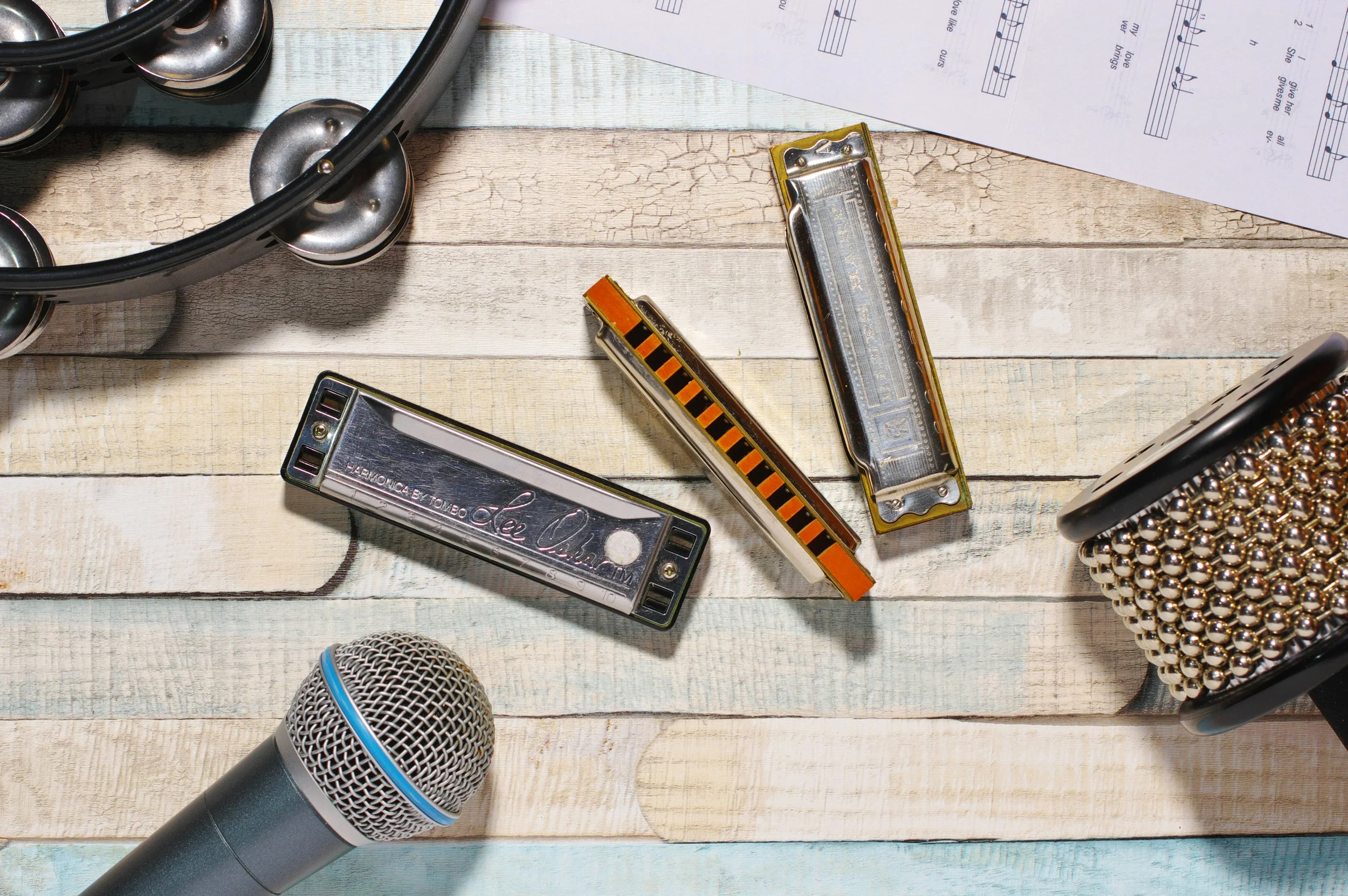 Close-up of musical instruments and a microphone on a wooden surface, including a harmonica, a small wooden block, a music sheet, and part of a tambourine.
