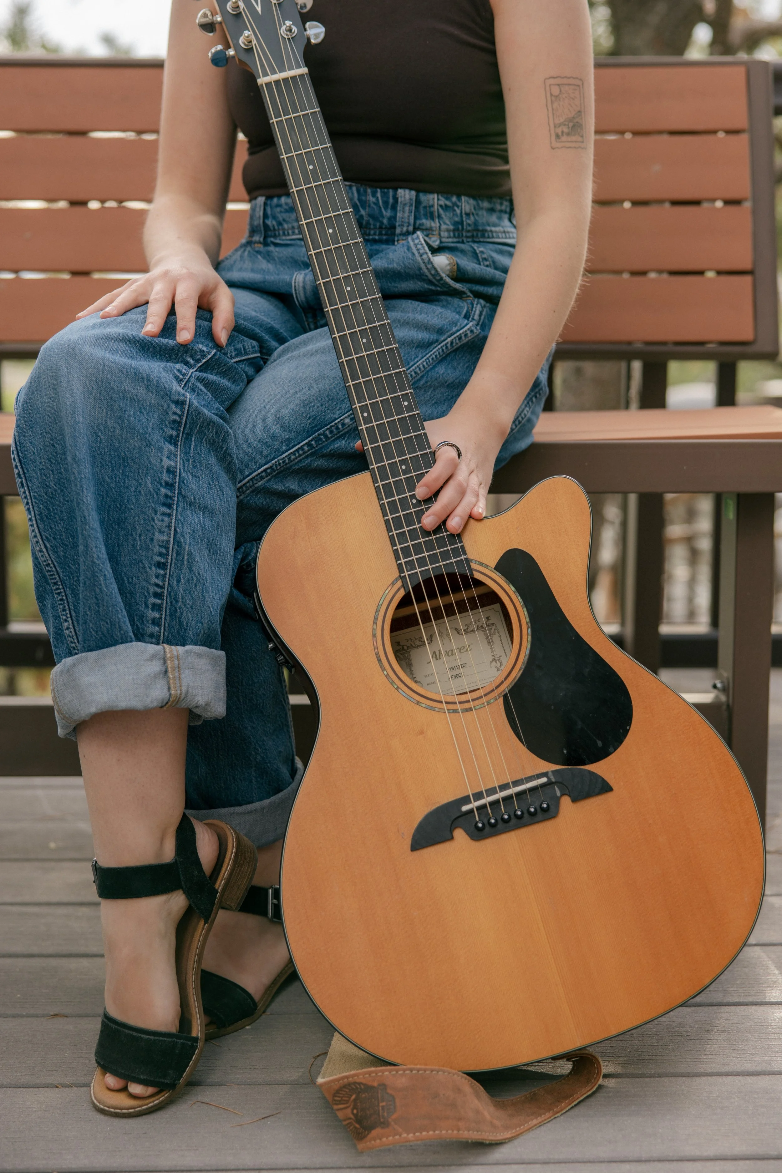 A person sitting on a wooden bench holding an acoustic guitar. They are wearing rolled-up jeans, a black shirt, and sandals, with a tattoo on their arm.